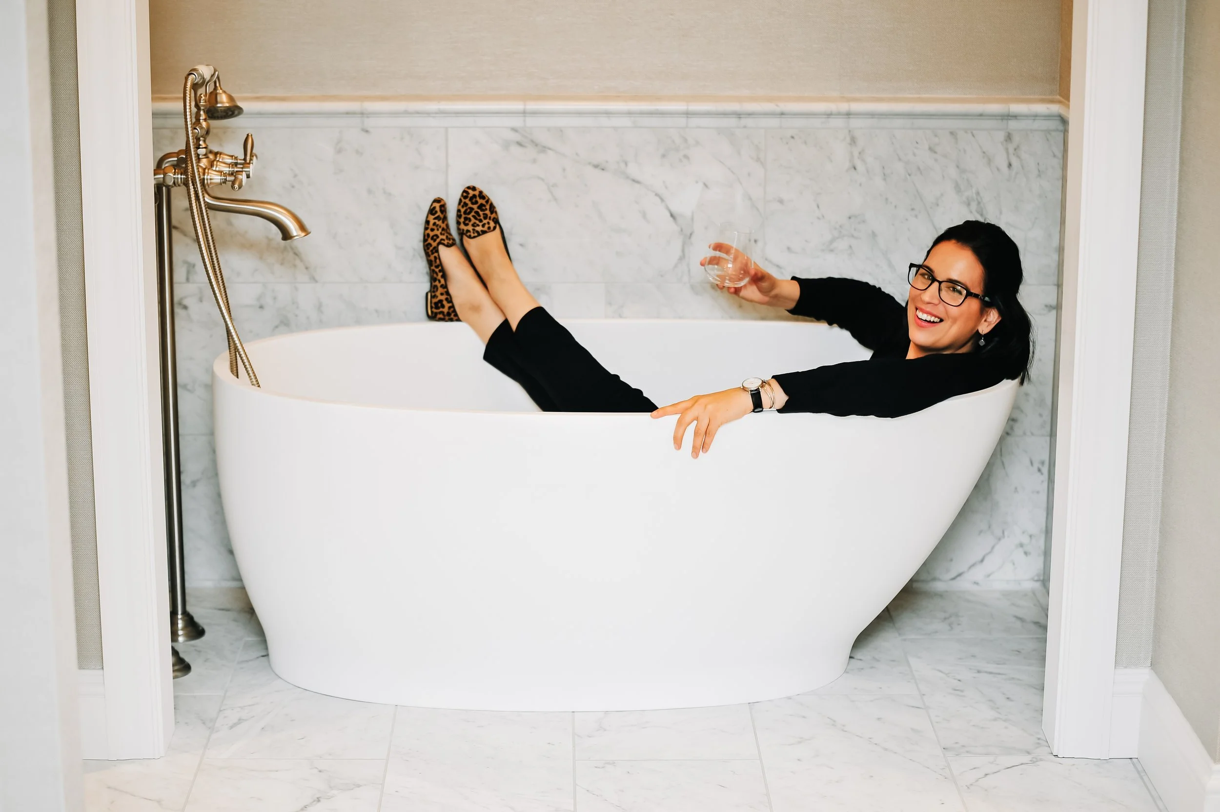 Woman relaxing in a white bathtub with leopard print shoes, wearing black clothing, glasses, and smiling, holding a glass of wine.