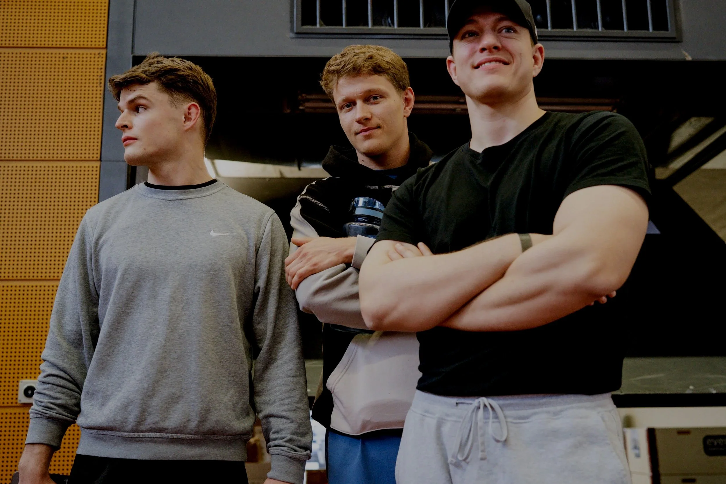 Three young men standing indoors, with two of them facing forward and one facing sideways. They are casually dressed and appear to be in a modern, well-lit environment.