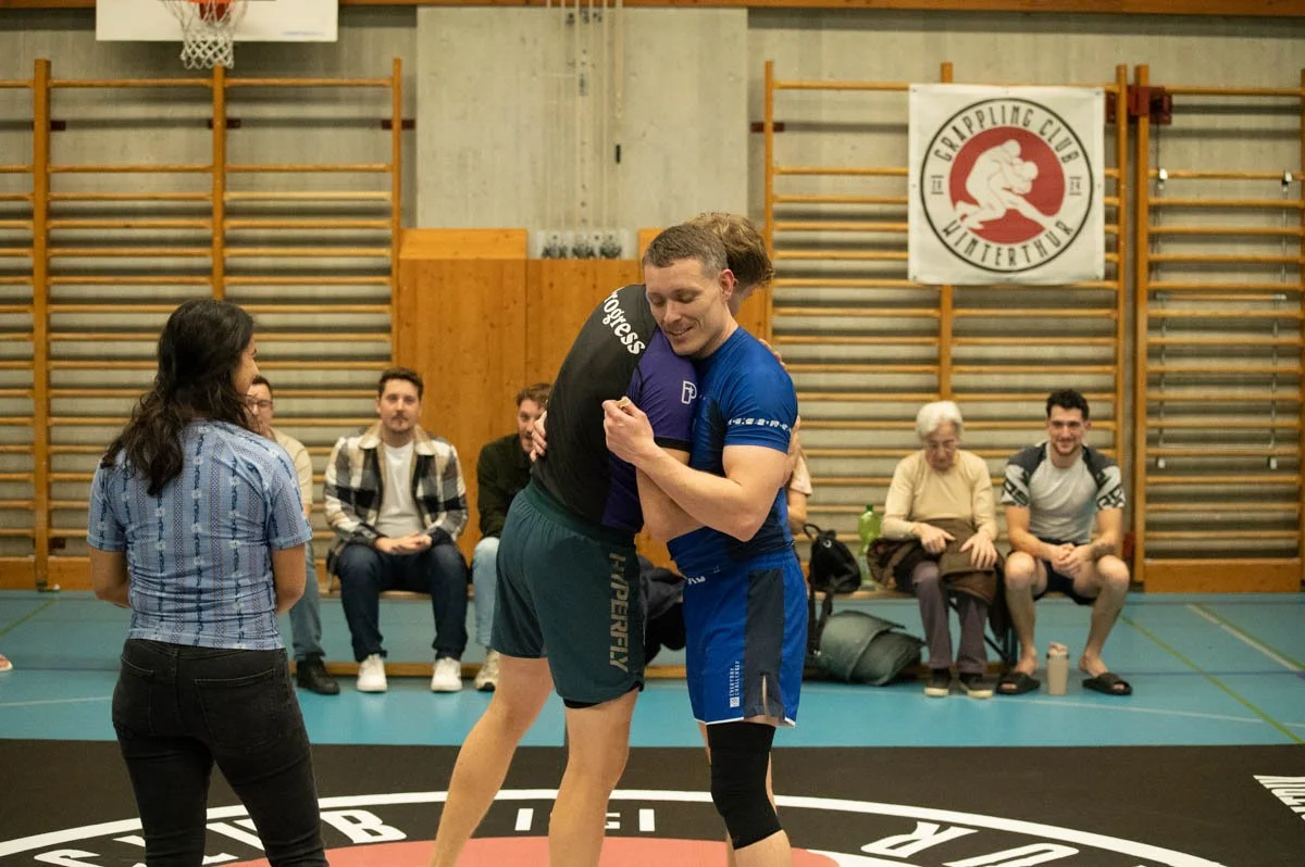 Two men hugging on a wrestling mat in a gymnasium, with spectators seated on benches and a logo for a grappling club on the wall behind them.