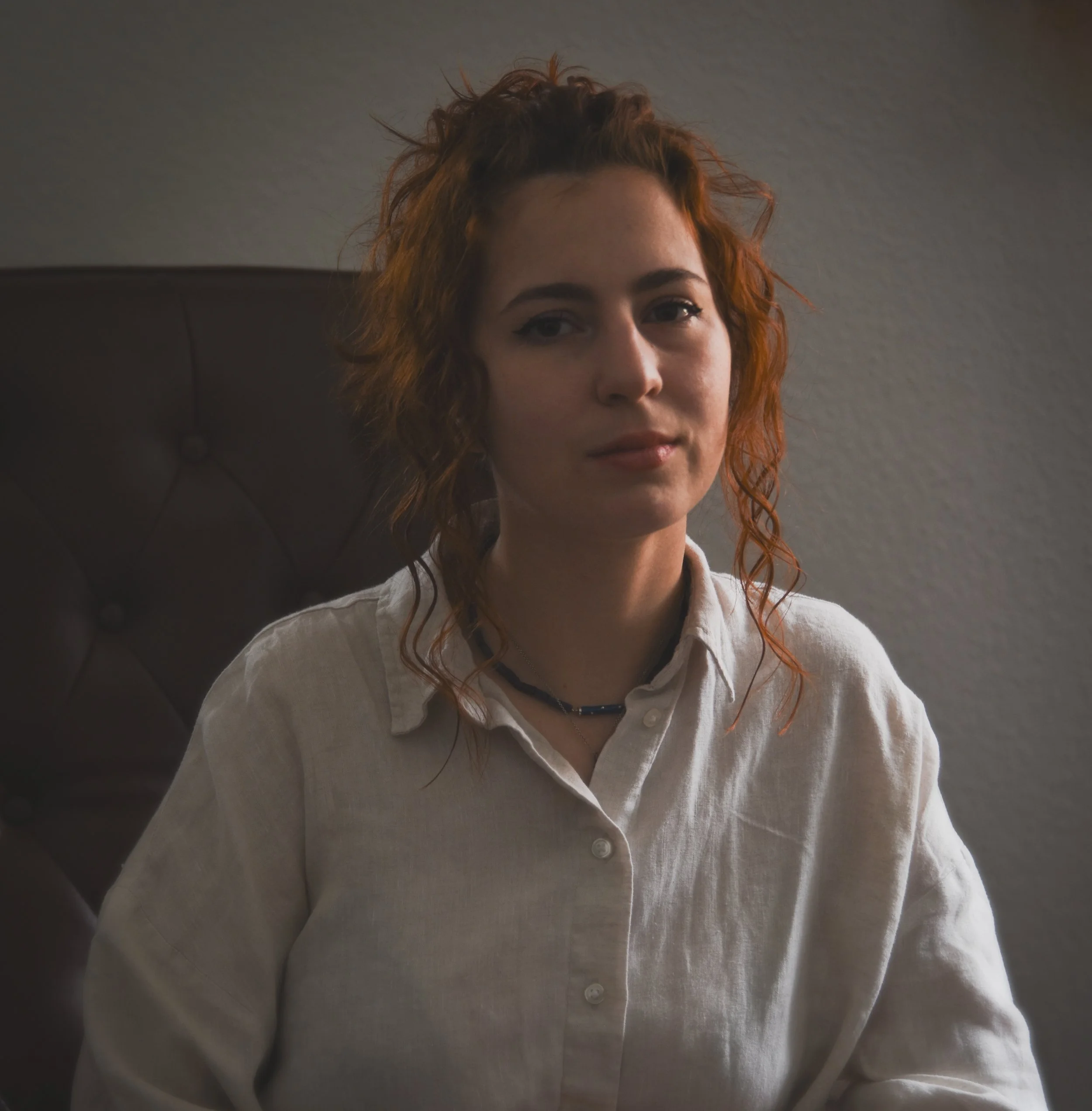 Close-up of a woman with red curly hair wearing a white button-up shirt and a dark necklace, sitting against a plain wall.