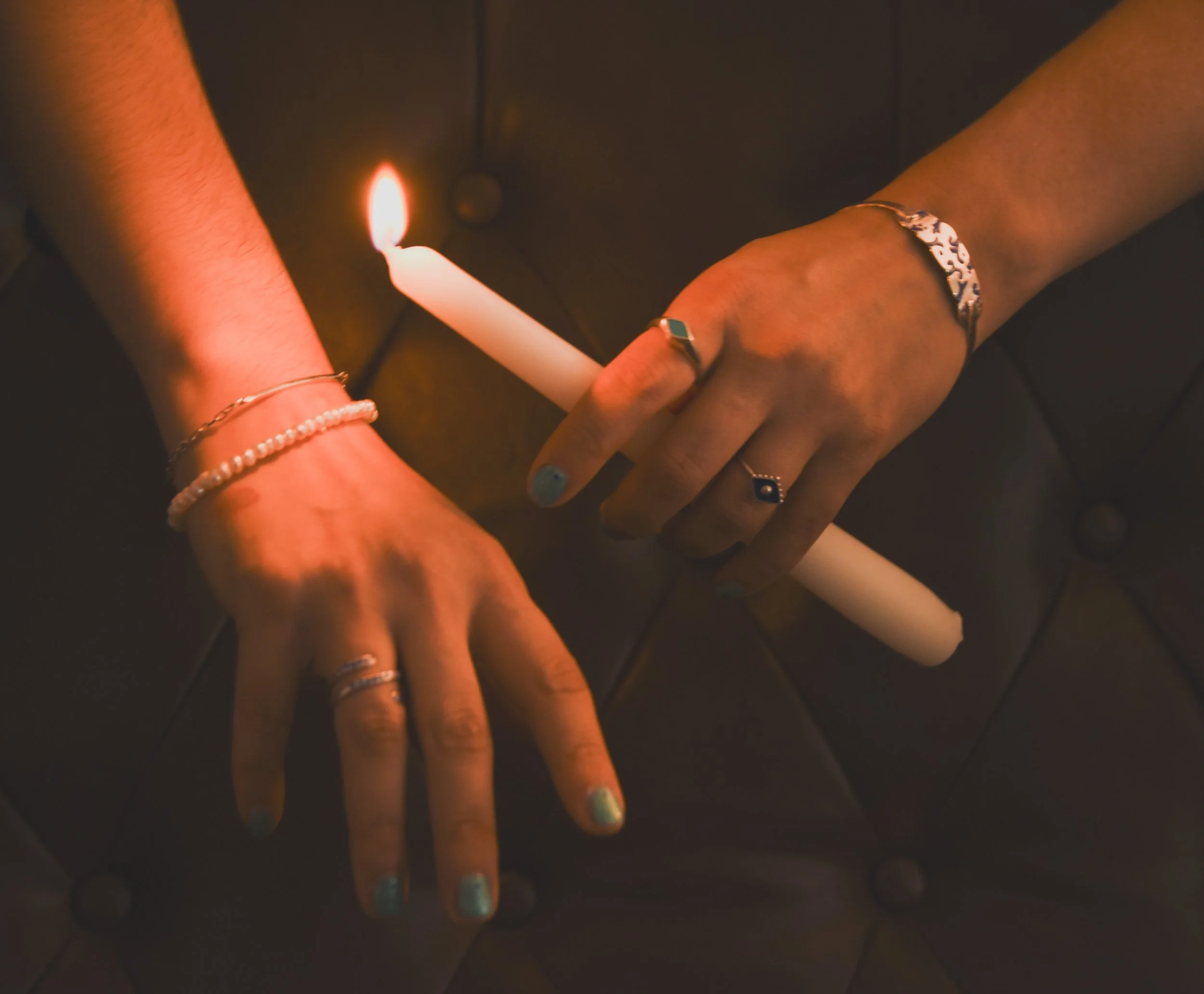Multiple hands with rings and bracelets are holding a lit white candle against a dark background.