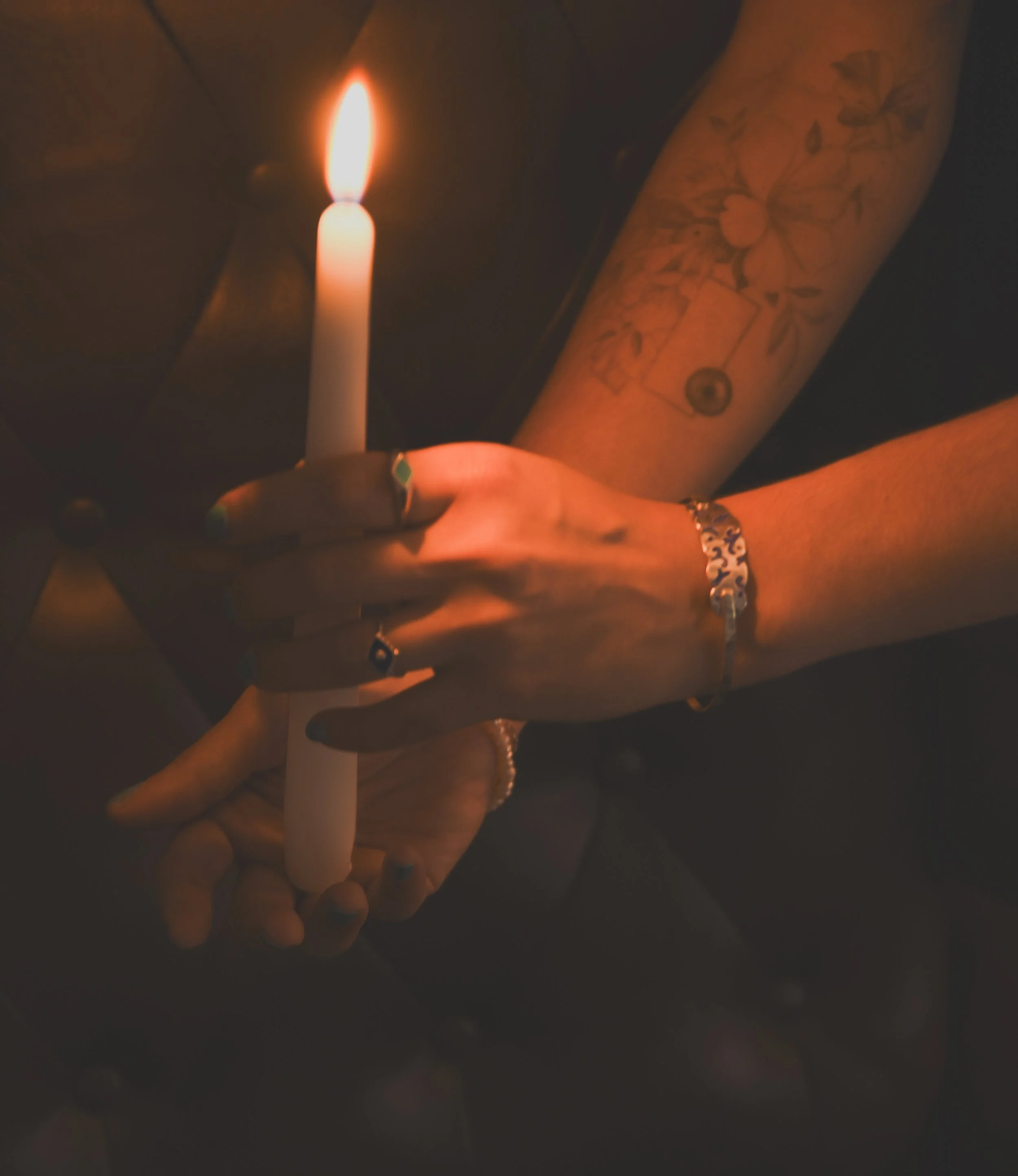 A hand holding a lit candle in a dark environment, with a visible tattooed arm and rings on the fingers.