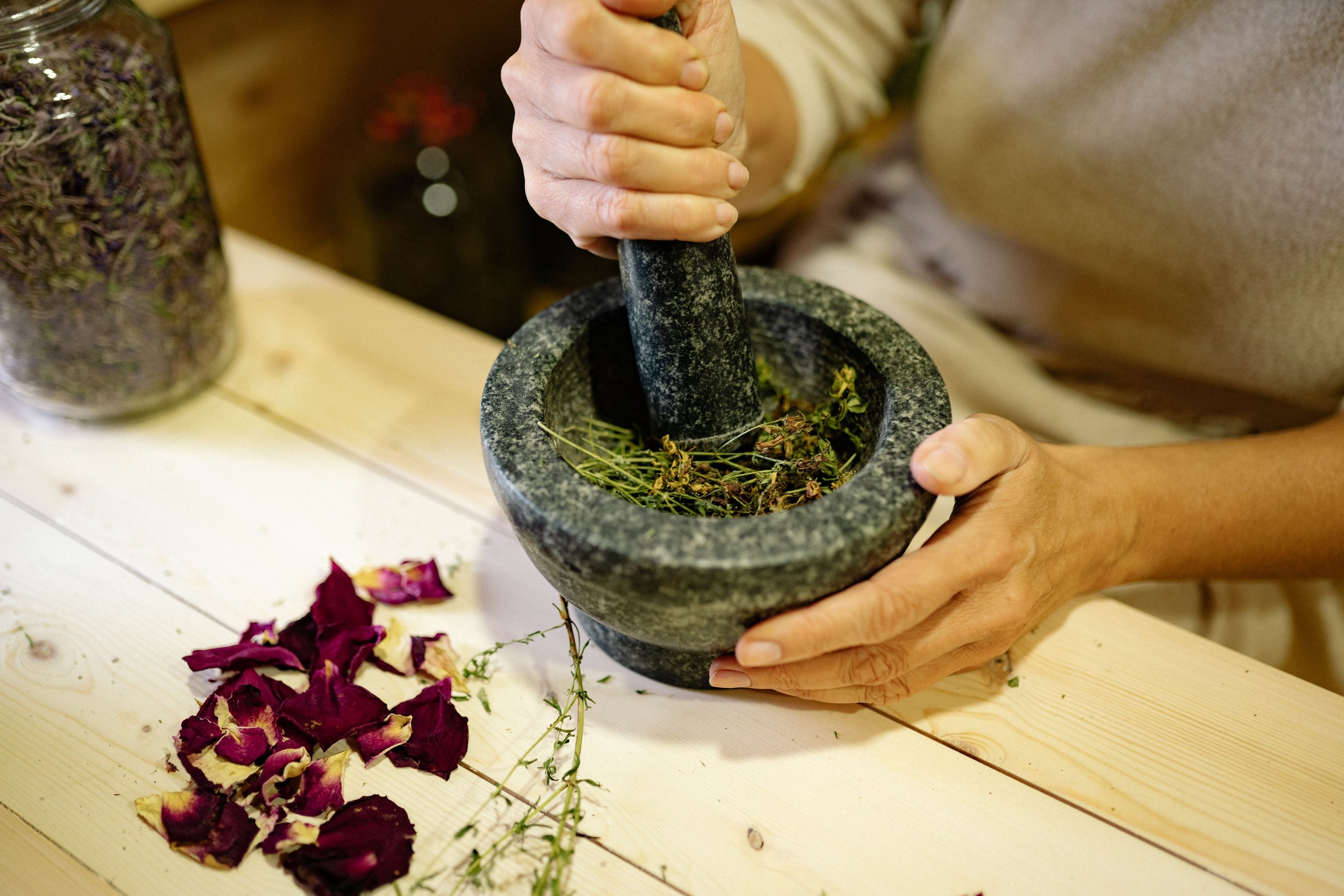 Person grinding dried herbs in a mortar and pestle on a white wooden surface, with dried rose petals scattered around.