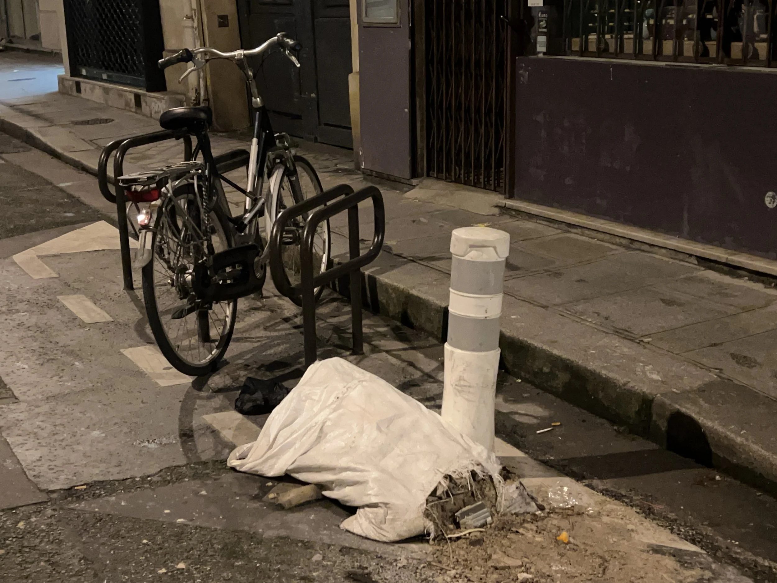A bicycle is locked to a bike rack on the sidewalk at night. There are bags or sacks on the ground near the bike, with some plastic debris and a small yellow object visible nearby. A pole or bollard is also in front of the bags. The background shows a closed gate and a dark building wall.