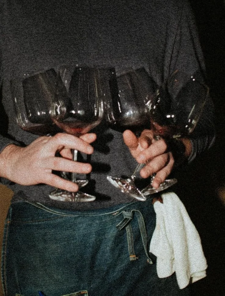 Person holding four glasses of red wine, wearing a dark sweater and jeans, with a white cloth tucked into the jeans pocket.
