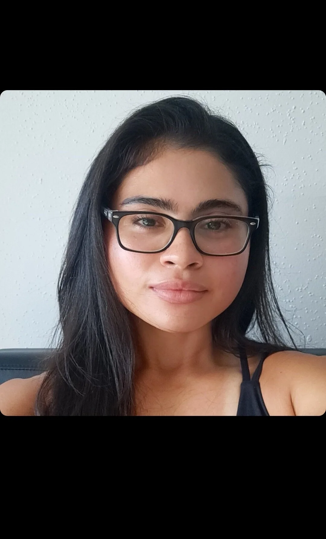 A young woman with long dark hair, glasses, and a neutral expression, wearing a black top, sitting against a white textured wall.