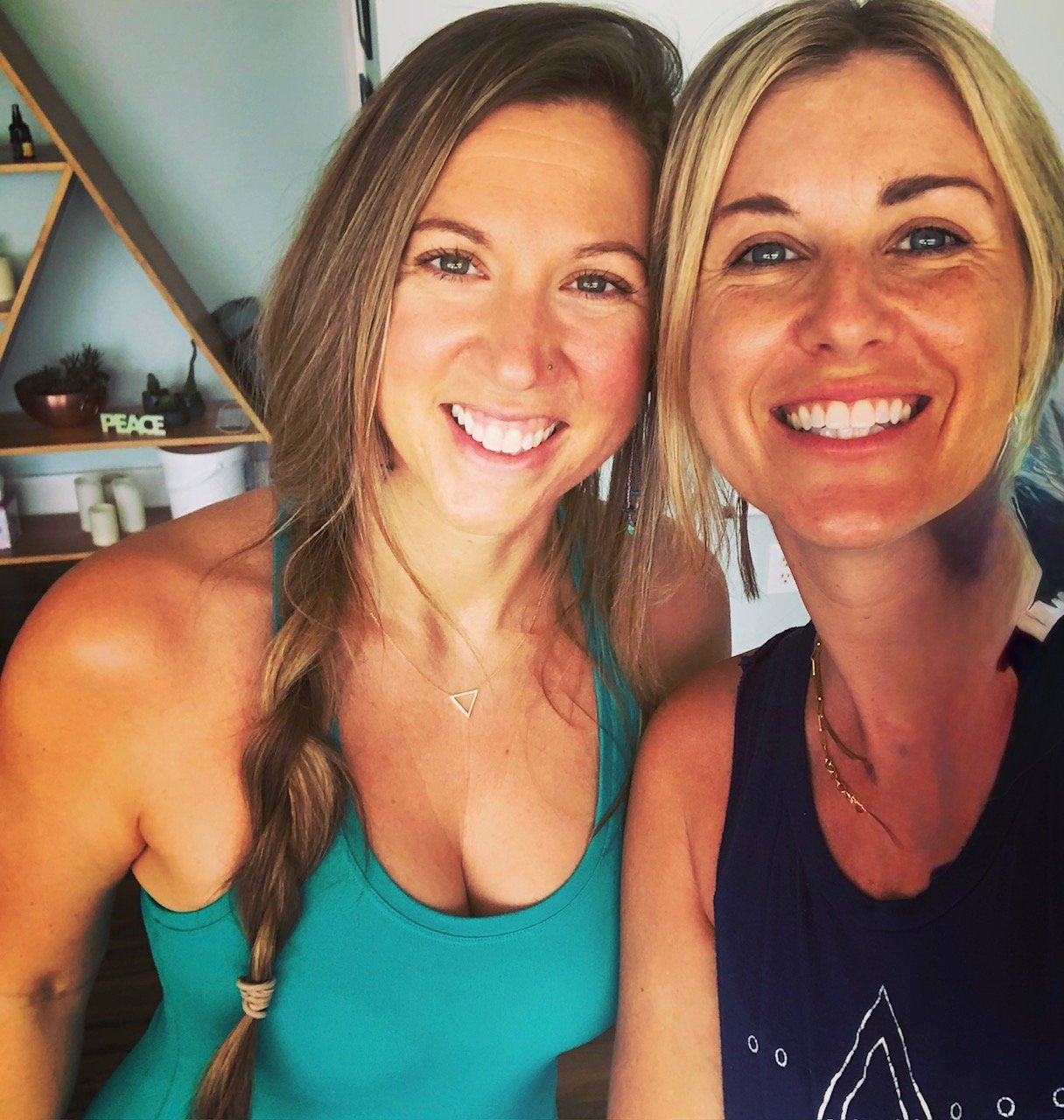 Two women smiling and taking a selfie together indoors. One has long, braided hair and is wearing a teal tank top, the other has short blonde hair and is wearing a dark sleeveless top. In the background, shelves with plants, candles, and decorative items are visible.