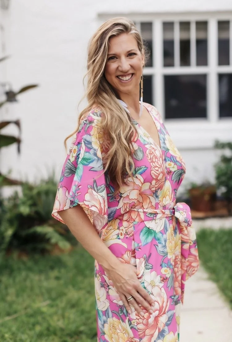 A woman with long, wavy blonde hair smiling outdoors, wearing a pink floral dress with a belt, in front of a white house with large windows.