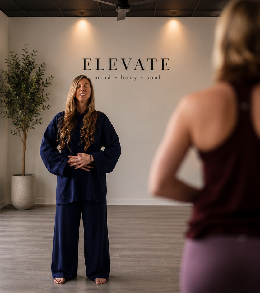 Two women in a yoga studio, one leading a class with eyes closed and hands in a meditation position, the other participant facing the instructor with her back to the camera. The studio has a sign that reads 'ELEVATE' and the words 'mind,' 'body,' and 'soul' underneath. There's a potted plant to the left.