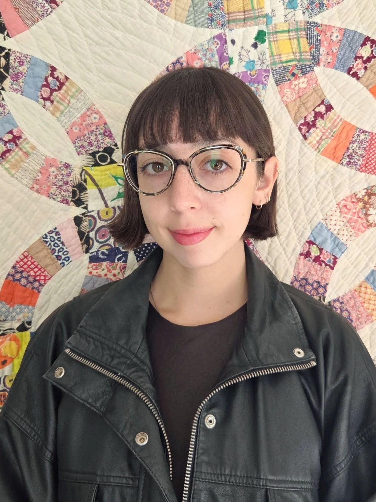 A young woman with short brown hair and glasses, wearing a black leather jacket, stands in front of a colorful quilt with floral and geometric patterns.