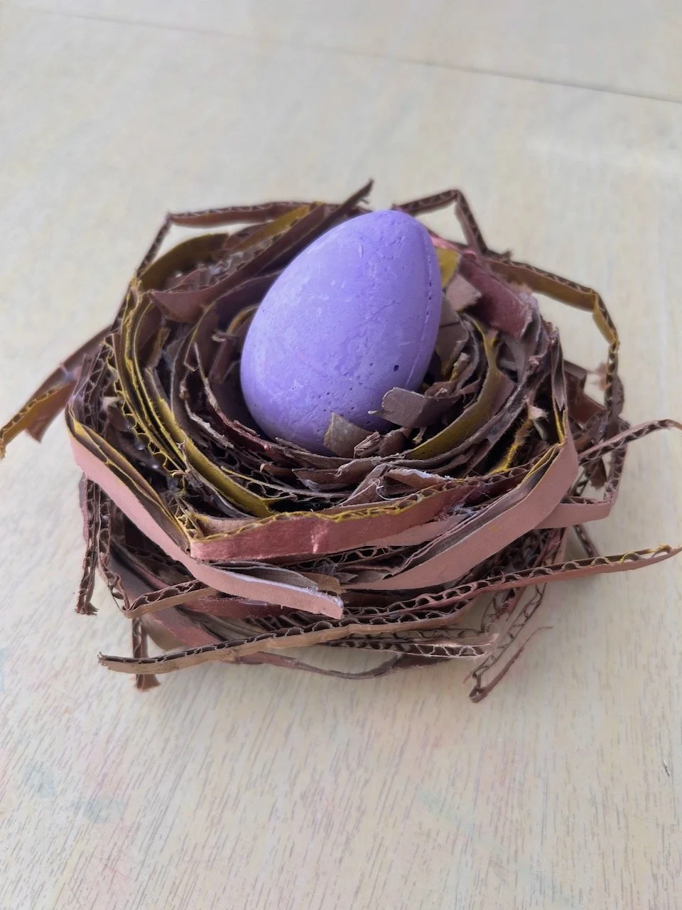 Purple egg in a nest made of layered, rolled cardboard pieces on a light-colored surface.