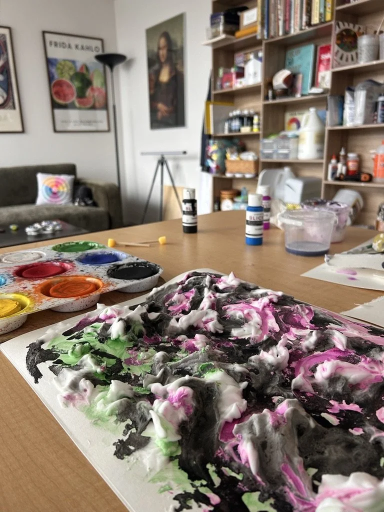 Abstract mixed media artwork with swirling black, pink, green, and white paint on a sheet of paper, set on a wooden table in an art studio.