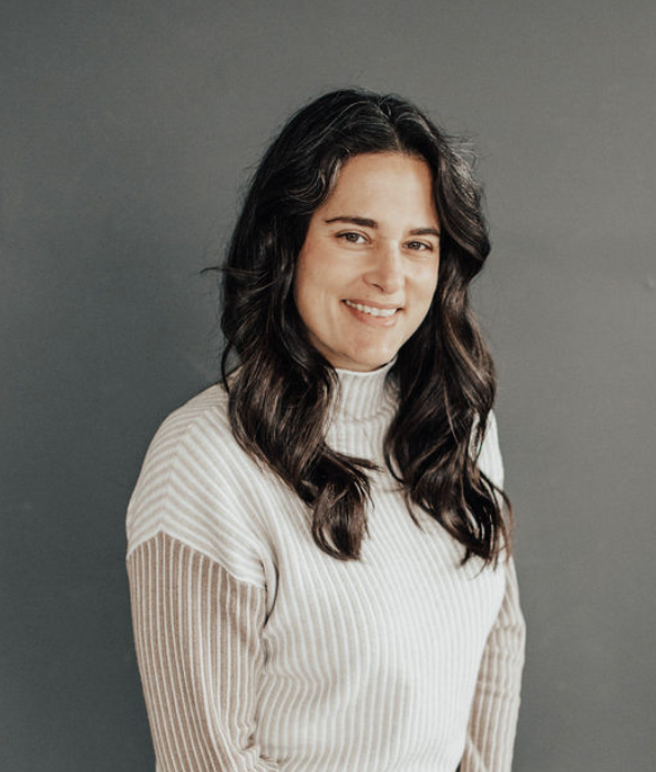 A woman with long dark wavy hair, smiling and wearing a cream-colored ribbed turtleneck sweater, standing against a plain gray background.