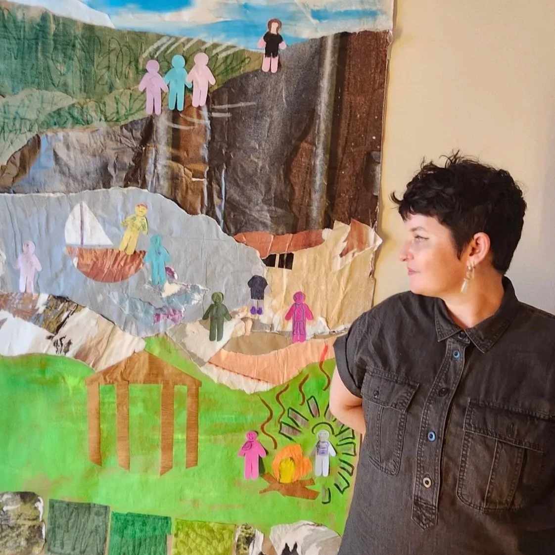 A woman with short dark hair and earrings stands next to a colorful handmade collage art piece with paper cutouts of children, landscapes, and a campfire scene.