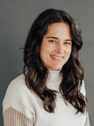 A woman with long dark hair, smiling, wearing a white, ribbed turtleneck sweater against a gray background.