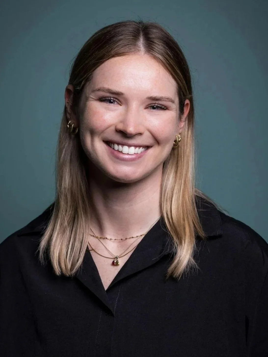 Portrait of a smiling woman with shoulder-length blonde hair, wearing gold jewelry and a black shirt, against a plain green background.