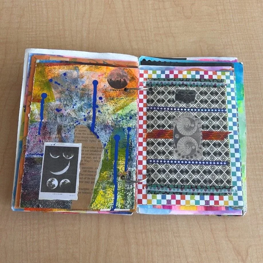 Open art journal with mixed media artwork, including colorful paint splashes, a black and white moon and planets sticker, and patterned paper elements.