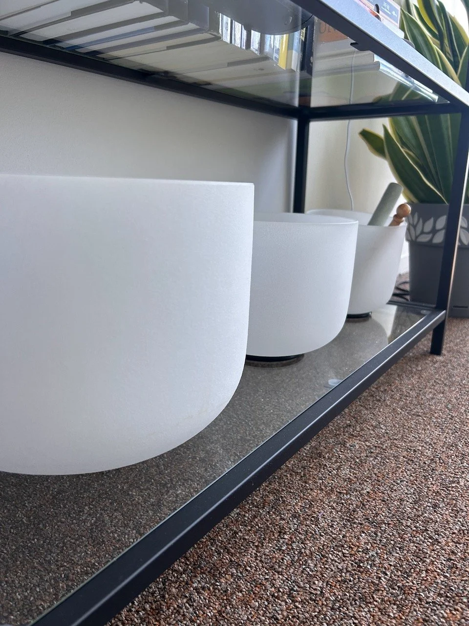 Close-up of a modern black metal table with glass top, displaying three crystal singing bowls and a tall potted plant with green leaves.
