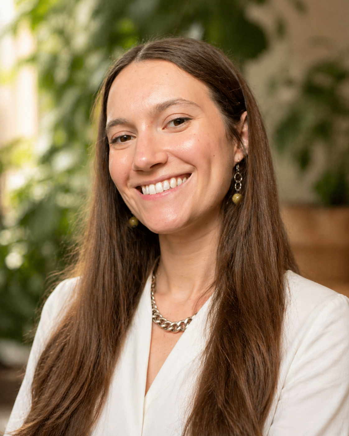 A woman with long brown hair smiling, wearing a white top, earrings, and a chunky necklace, standing in front of a blurred green background.