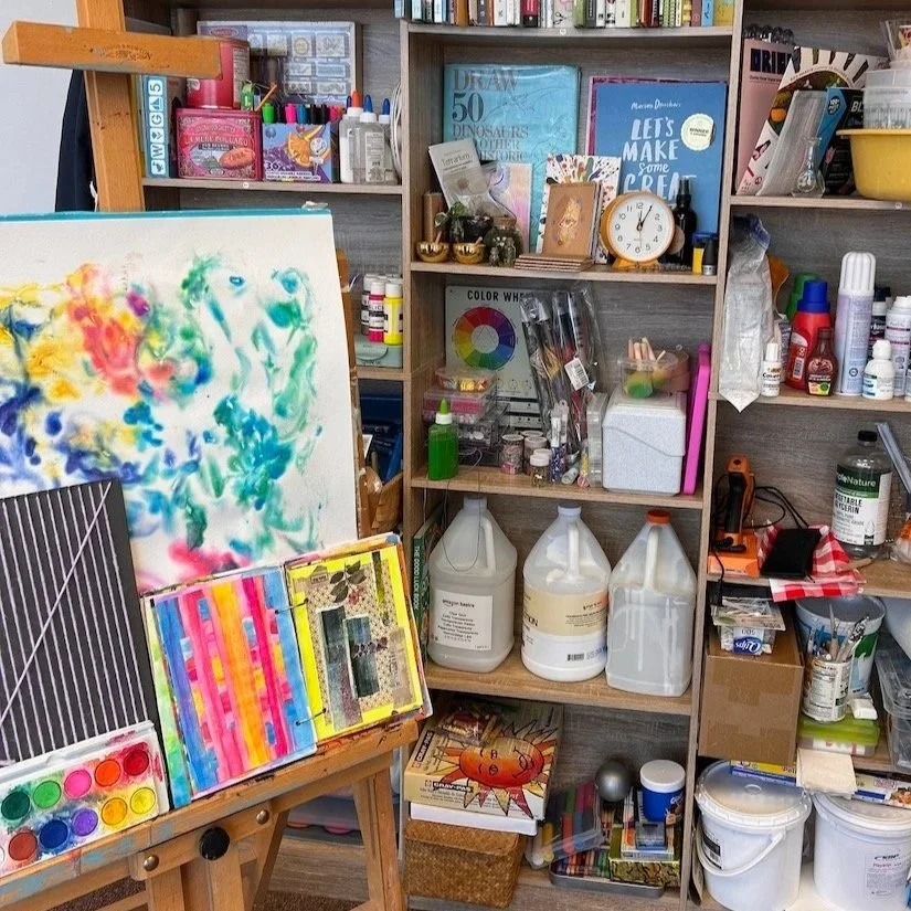An art studio workspace with shelves of art supplies, paints, brushes, and books, and an easel holding a colorful abstract acrylic painting.