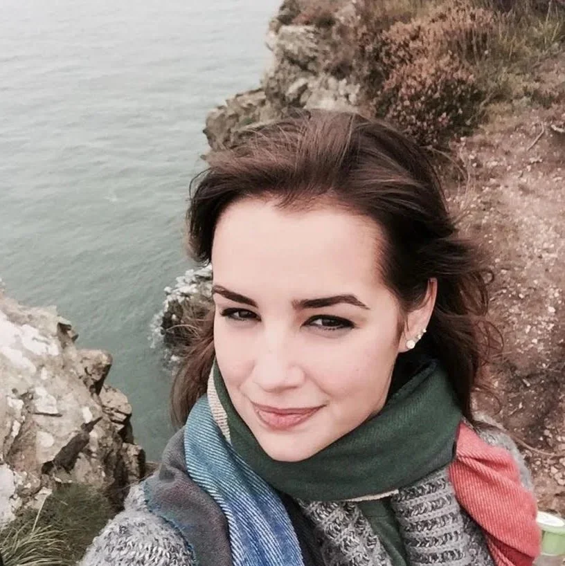 A young woman with brown hair, wearing a green scarf and a gray sweater, taking a selfie on a rocky cliffside above the ocean.