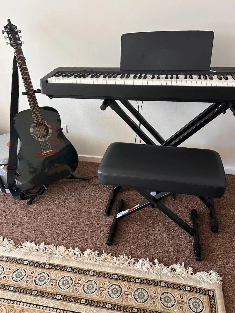 A black acoustic guitar resting on a stand, a digital keyboard on a stand, a black padded bench in front, all on a brown carpet with a decorative rug in the foreground.