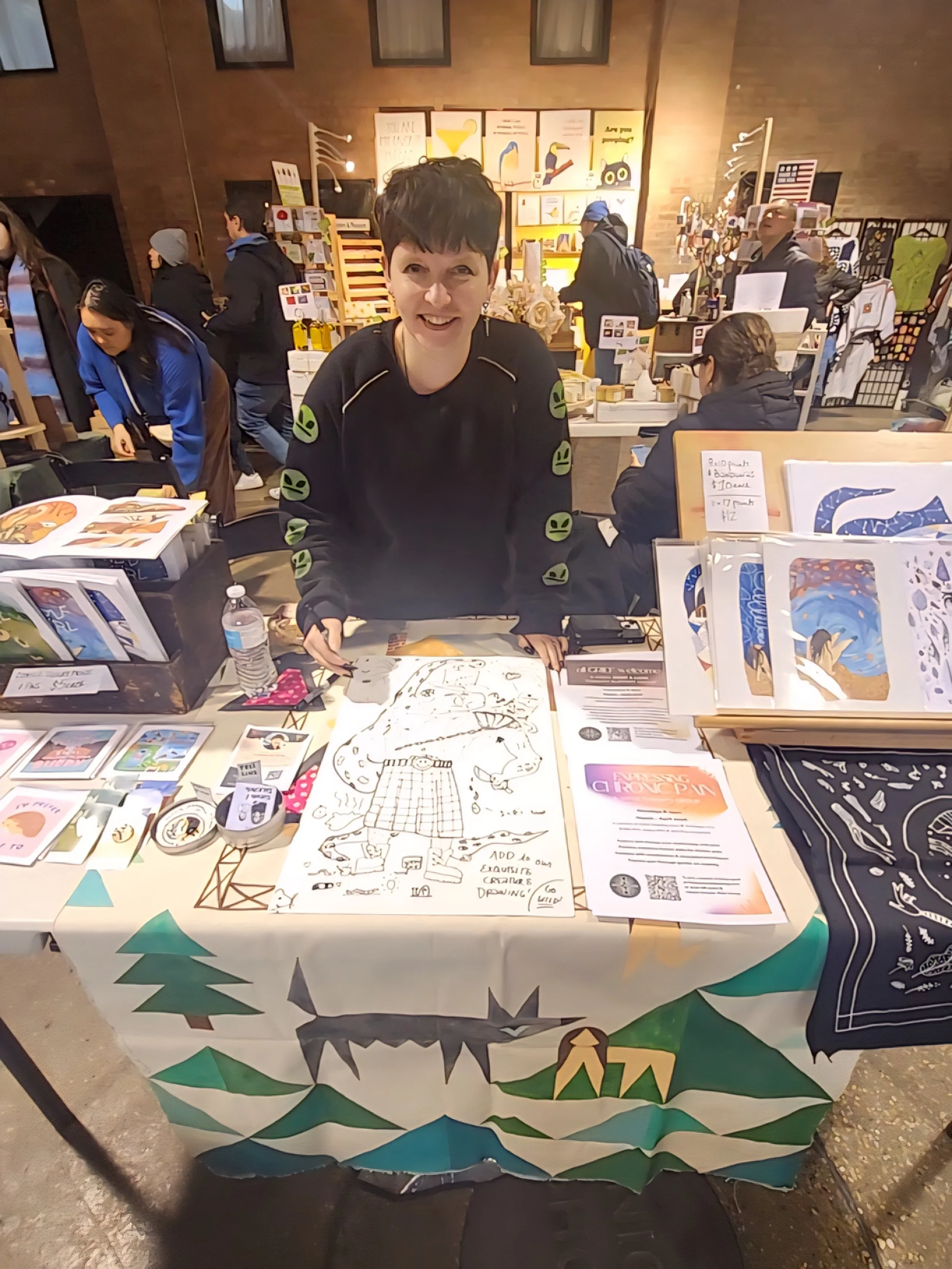 A nonbinary person behind a table at a craft fair, displaying artwork and prints, with other vendors and shoppers in the background.