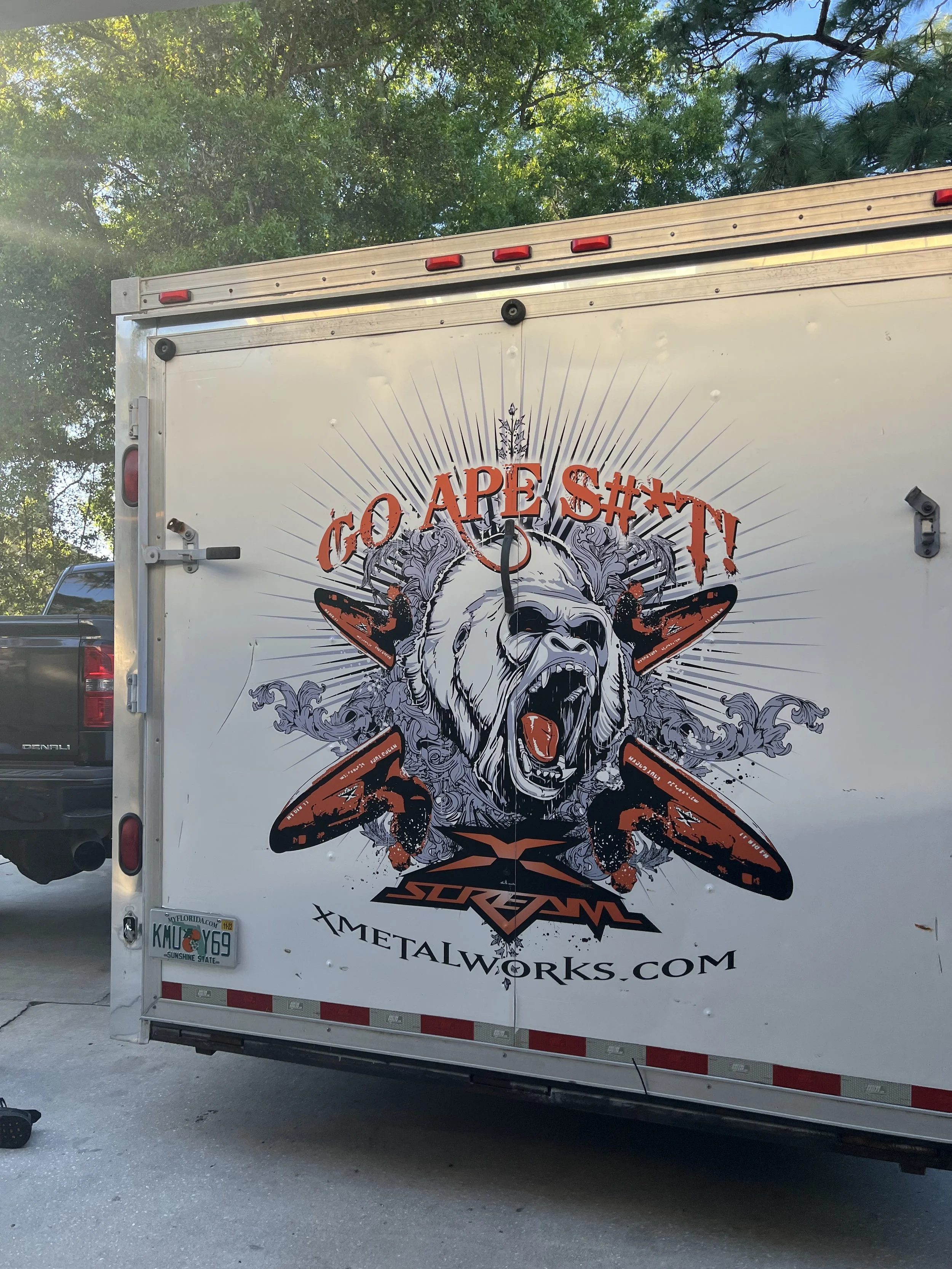 The back of a white trailer with a graphic of a roaring bear with wings, holding a tattoo machine, and the words 'GO APE SH#T!', with the website 'XMETALWORKS.COM' below.