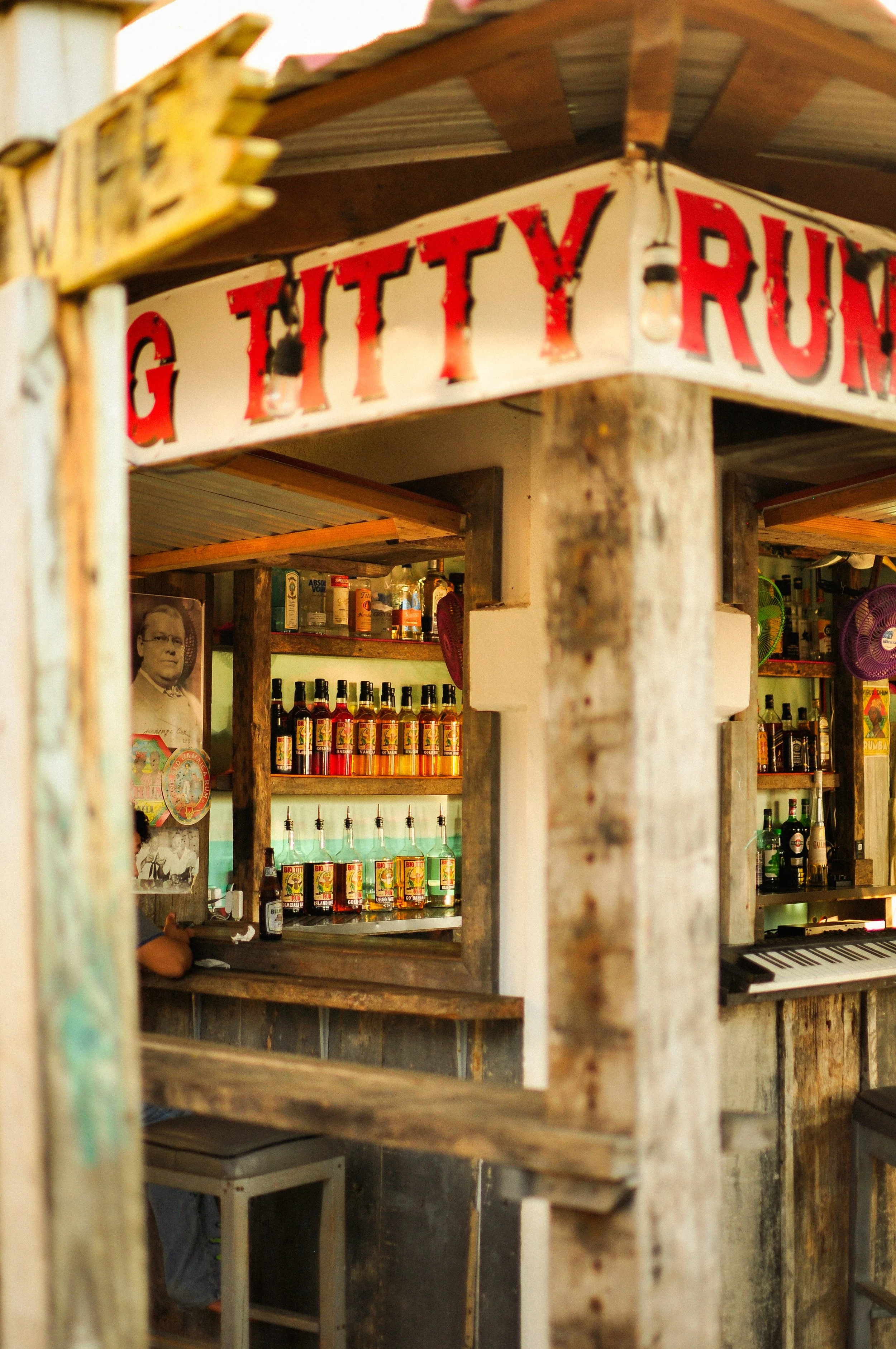 A rustic bar with a red and white sign that says 'GITTY RUM'. The bar has bottles of alcohol and a keyboard, with a wooden structure and corrugated metal roof.