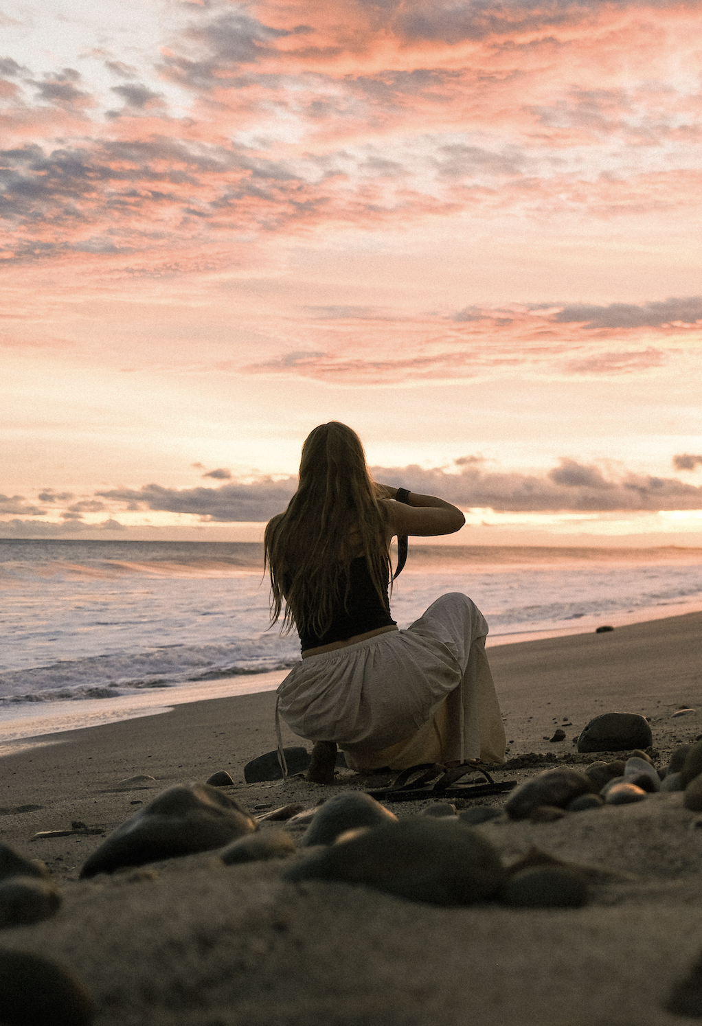 A woman with long hair, sitting on the sand at the beach, watching the sunset with a camera in hand.