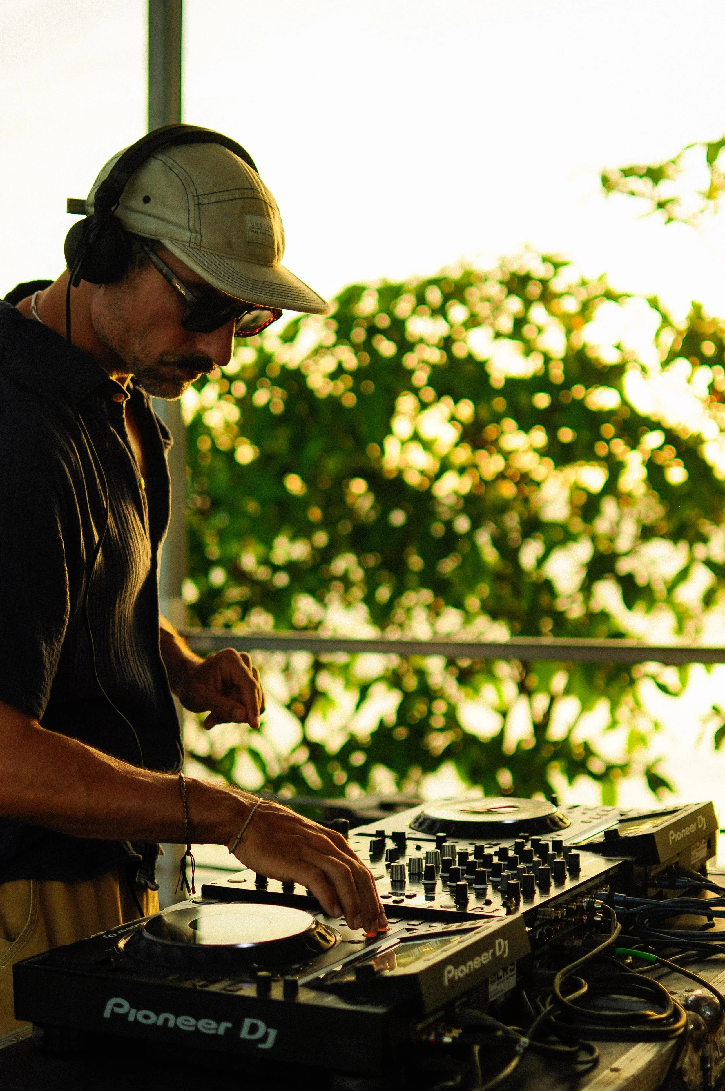 A DJ wearing a beige cap, black headphones, and sunglasses is playing music on a Pioneer DJ controller outdoors during the daytime, with trees in the background.