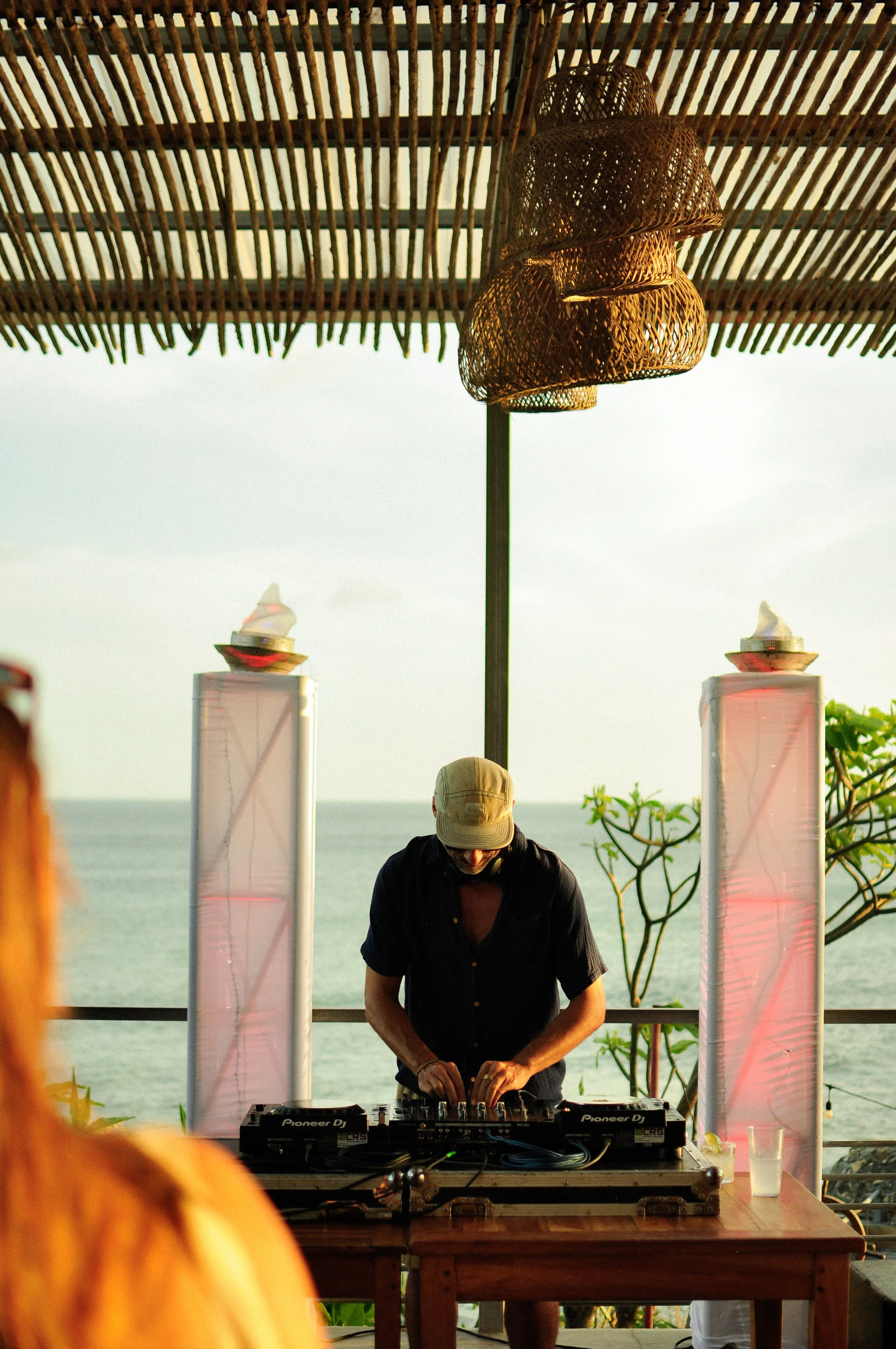 A DJ wearing a beige hat and dark shirt is DJing at an outdoor event during the daytime, with a view of the ocean in the background. The DJ is using Pioneer DJ equipment. A wicker lampshade hangs overhead, and there are two tall, pink-tinted transparent panels with candles or small lights inside. The setup is under a bamboo or wooden canopy, and a tree with green leaves is visible on the right side.