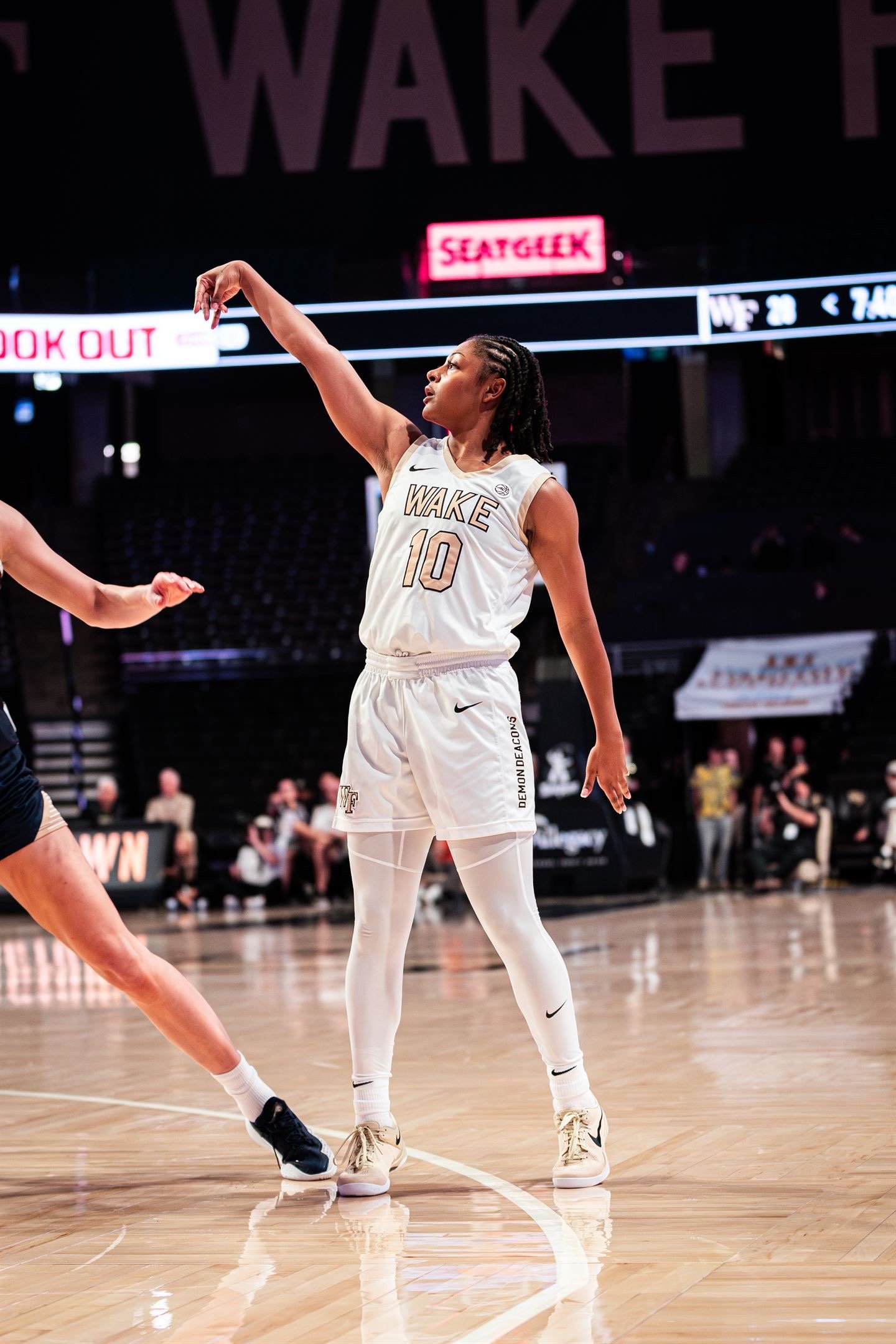 A female basketball player with braided hair in a white uniform with the number 10, reaching upwards during a game on a basketball court.