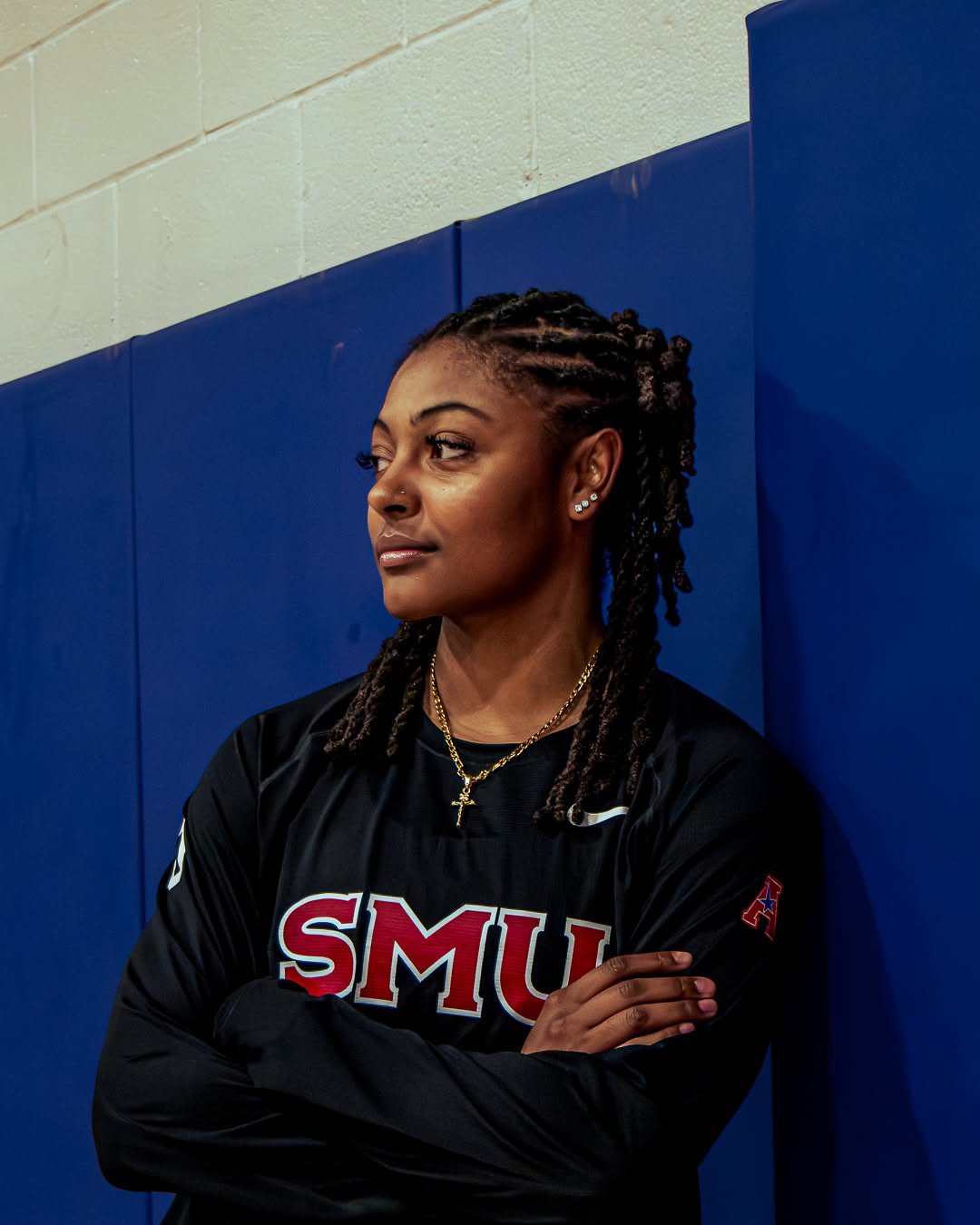 A woman with braided hair wearing a black sports jacket with SMU in red and white letters, standing against a blue padded wall with arms crossed.