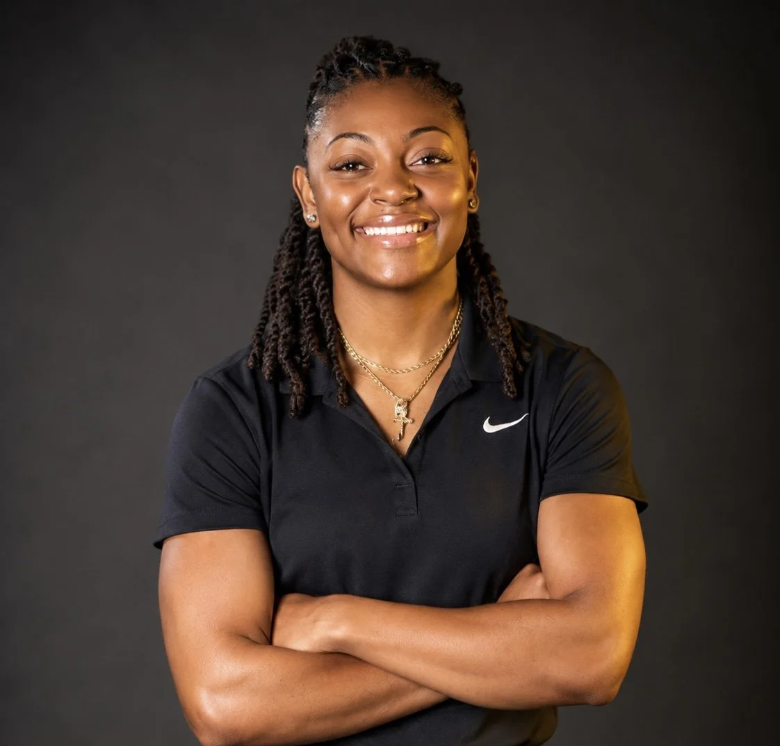 A smiling woman with braided hair wearing a black Nike polo shirt and layered gold necklaces against a dark background.