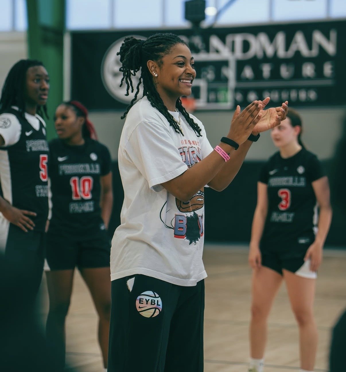A group of young women on a basketball court, with some clapping and smiling. They are wearing sports uniforms and are standing in front of a green wall with a sign that says 'Handyman's Nature'.