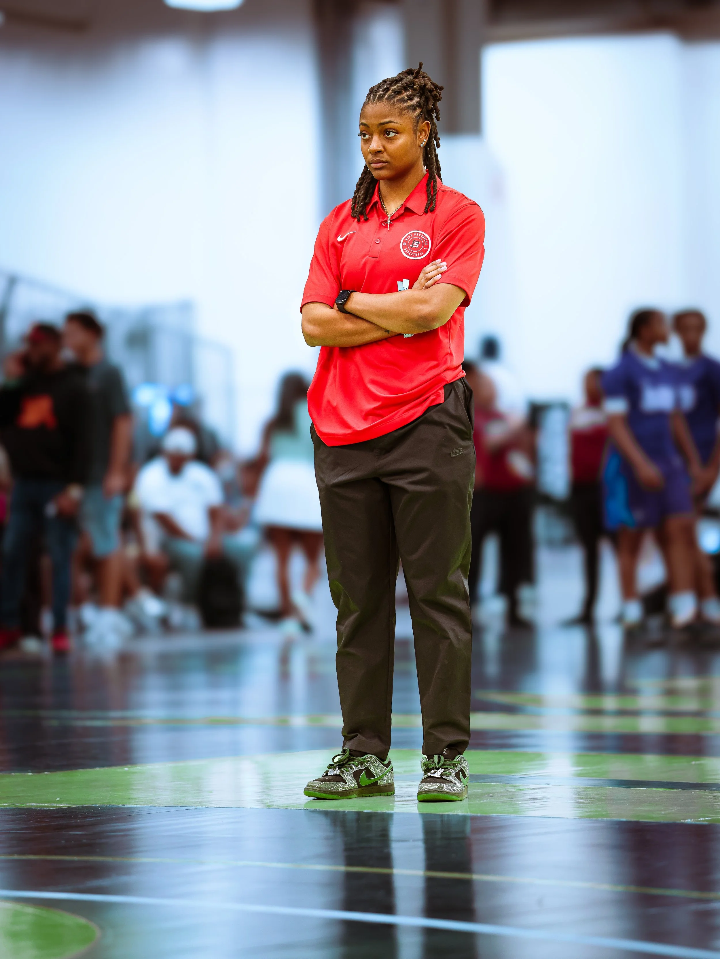 A young woman with dreadlocks stands with arms crossed, wearing a red sports polo, dark pants, and Nike sneakers inside a gymnasium with other people blurred in the background.