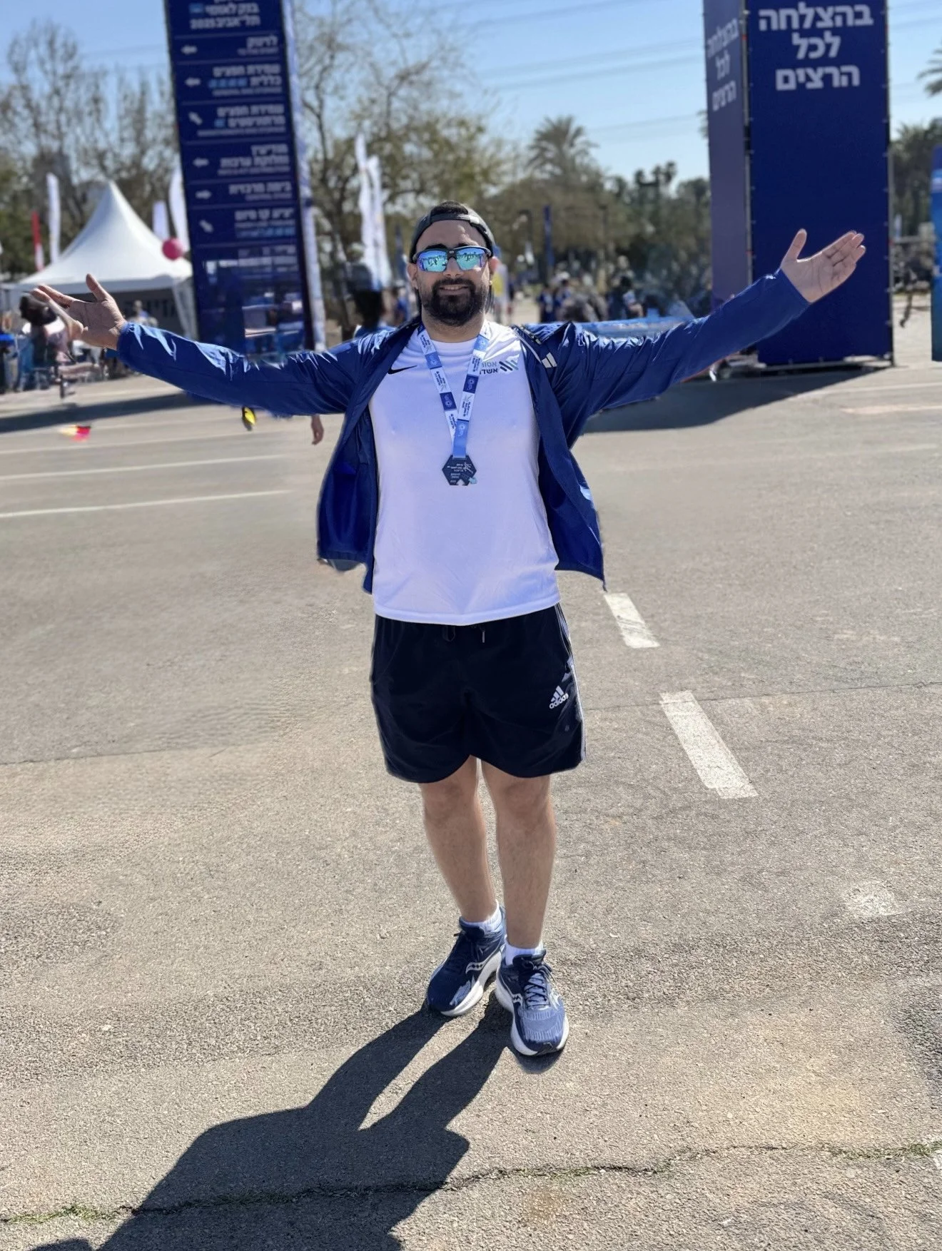 A man standing on a street with his arms outstretched, wearing sunglasses, a white shirt, black shorts, sneakers, and a medal around his neck at a race event with blue banners in the background.