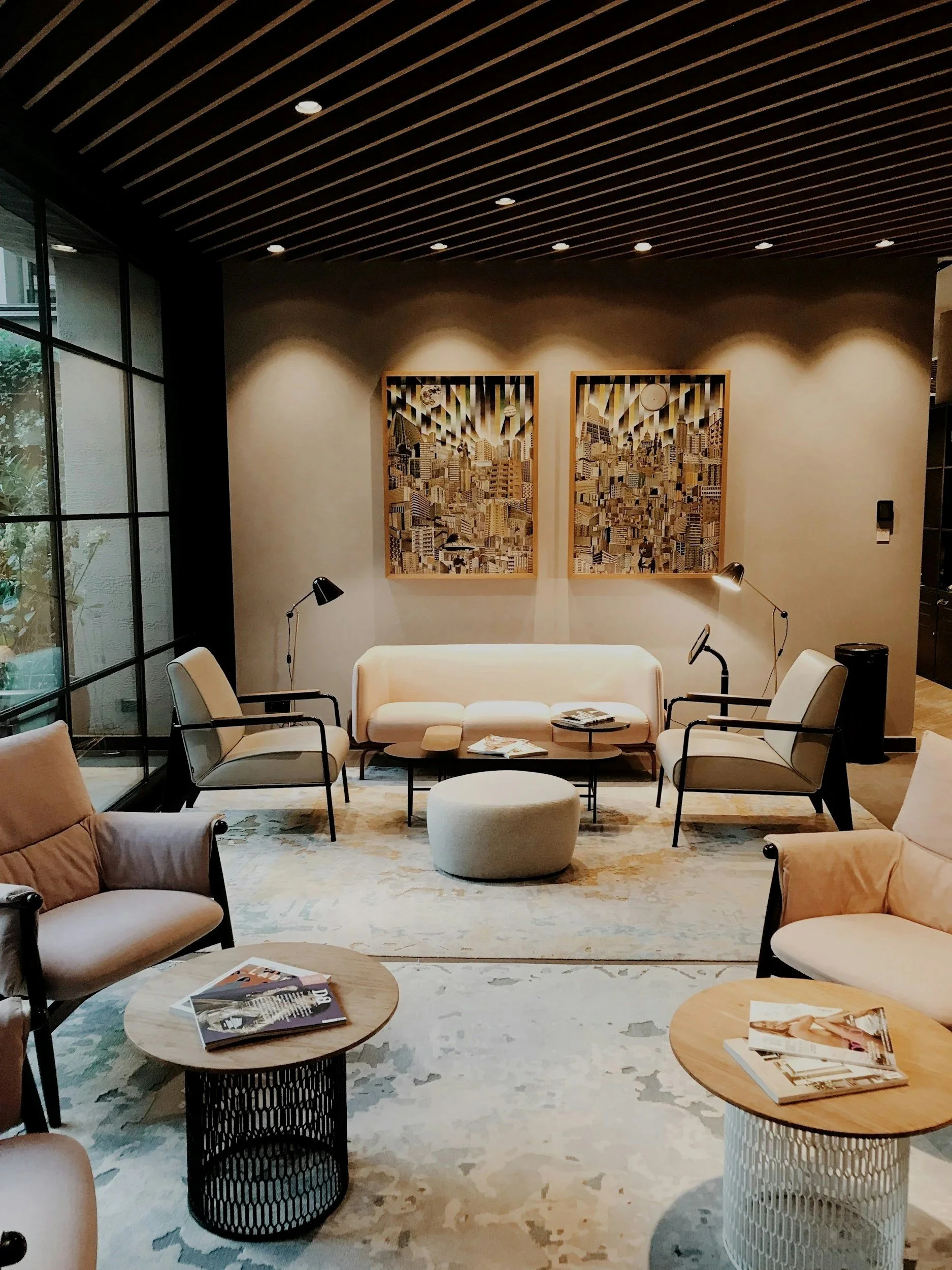 A modern, cozy lounge area with beige sofas, armchairs, and round coffee tables arranged on a patterned rug. Wall art featuring cityscapes hangs on the wall, illuminated by ceiling spotlights. A large glass window with a black metal frame is on the side, allowing natural light to enter.