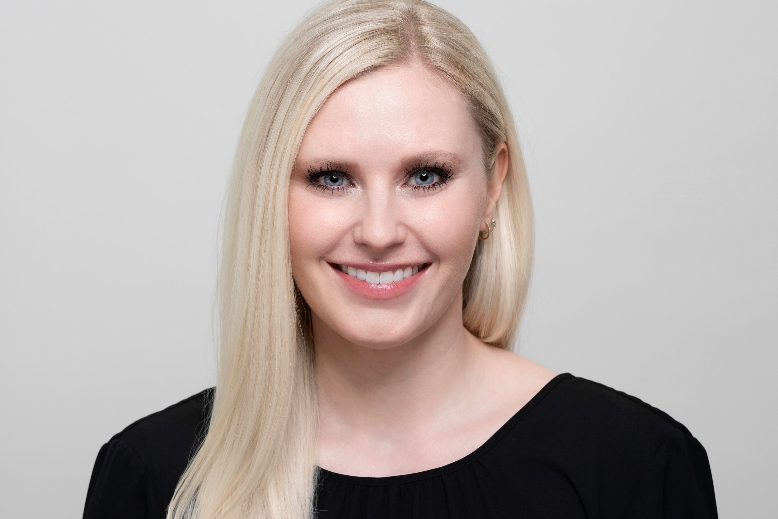 A young woman with long blonde hair, blue eyes, and a friendly smile, wearing a black top, against a plain light gray background.