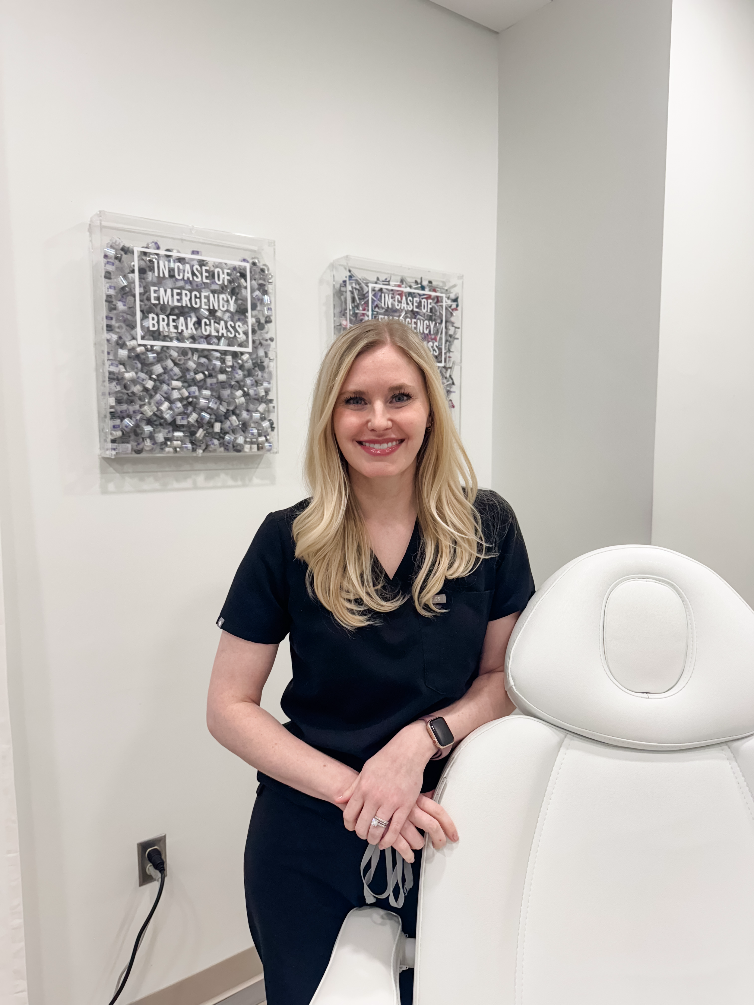 A woman in black scrubs wearing a smartwatch, standing next to a white massage chair, smiling in a medical or spa setting.