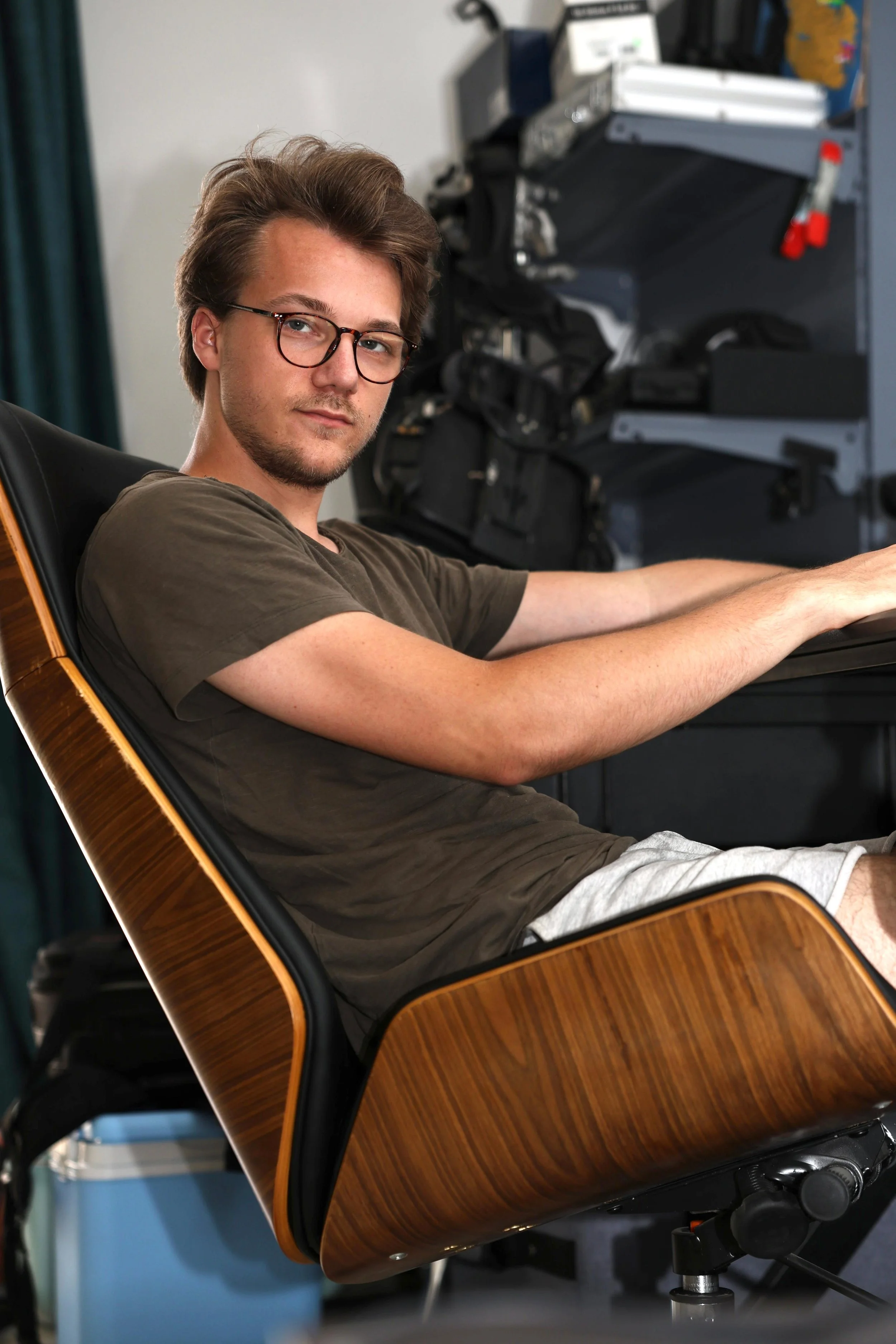 Junger Mann mit Brille sitzt entspannt in einem modernen Holzstuhl, im Hintergrund ist technische Ausrüstung sichtbar.