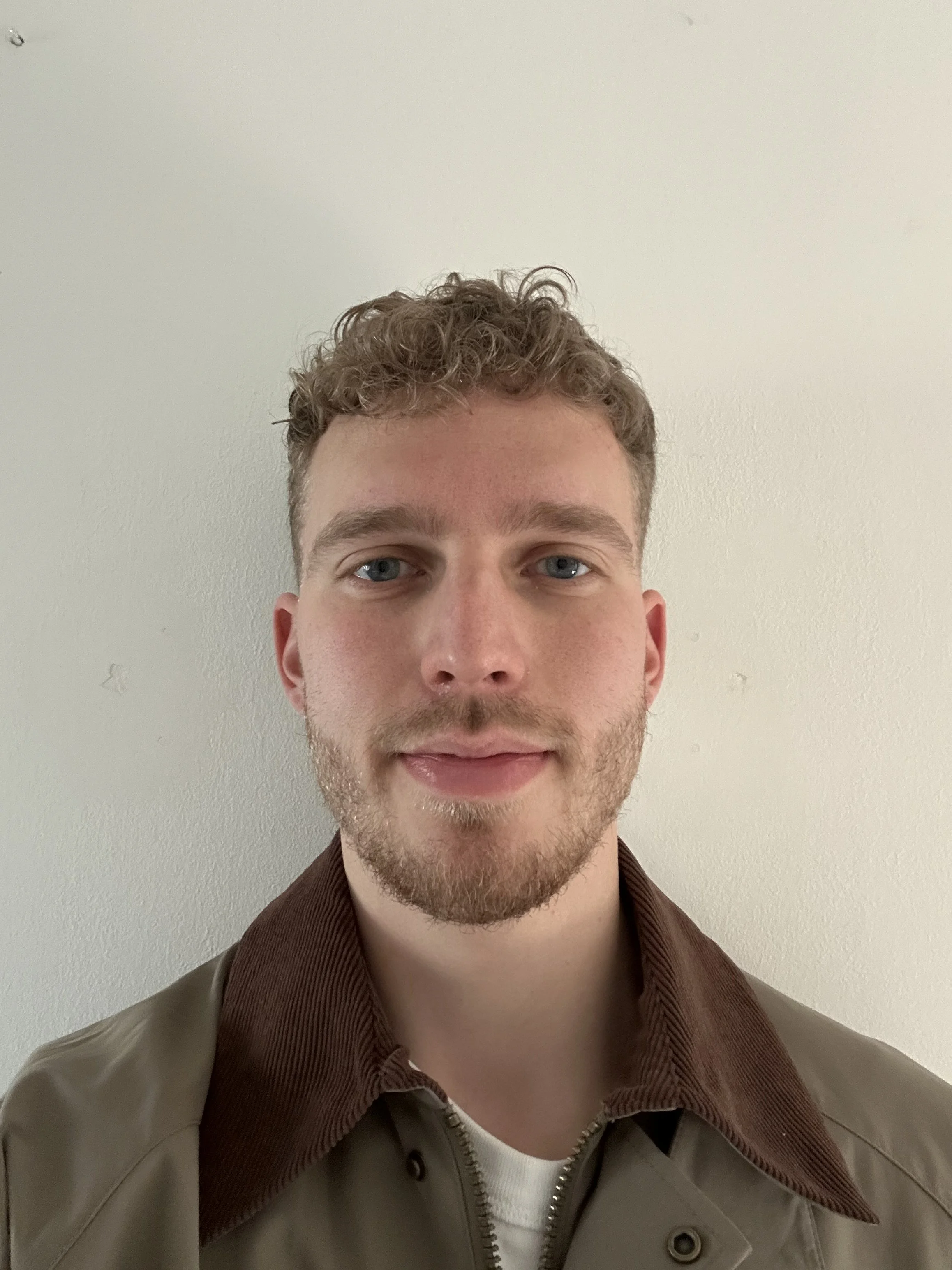 Portret van een jonge man met lichte huid, blauwe ogen, kort gekruld bruin haar en een lichte baard, gekleed in een bruin jack en wit T-shirt, staand voor een witte muur.