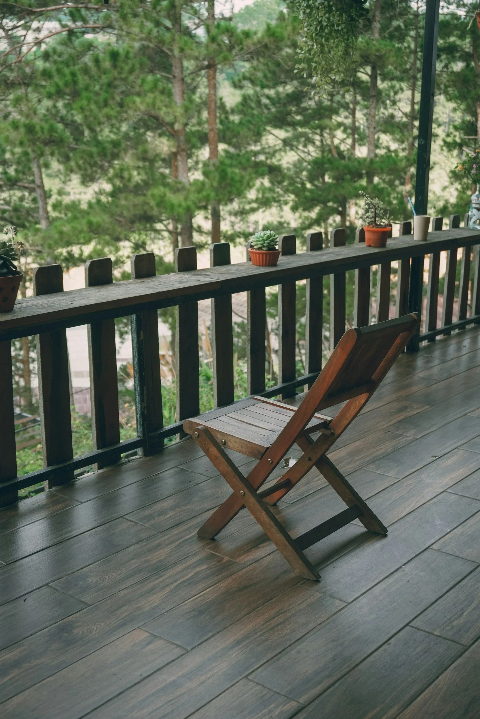 A balcony with a wooden folding chair, four potted plants, and a railing overlooking a forest of tall green trees.