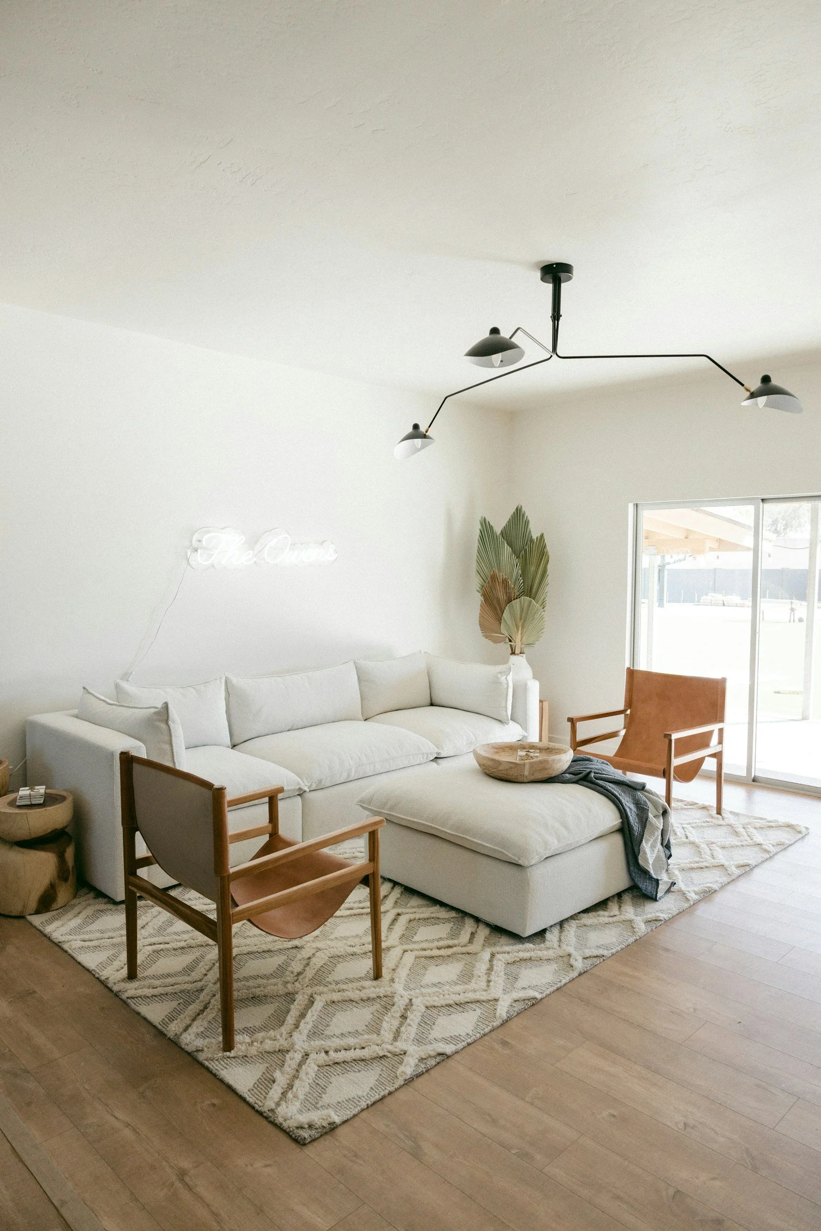 Living room with a white sectional sofa, wooden chairs, a patterned rug, and a large window. There is a neon sign on the wall that says 'The Oasis' and a decorative plant in a white vase. A black and white modern ceiling light fixture hangs from the ceiling.