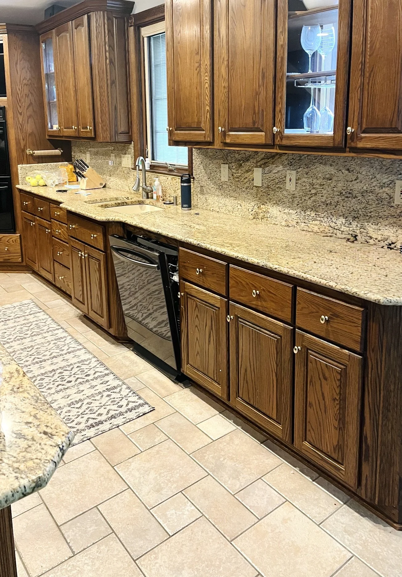Kitchen with wooden cabinets, granite countertop, a stainless steel sink, and a black dishwasher. There are glassware displayed in a cabinet, a window above the sink, and various kitchen items on the counter. The floor is tiled and there is a patterned rug.