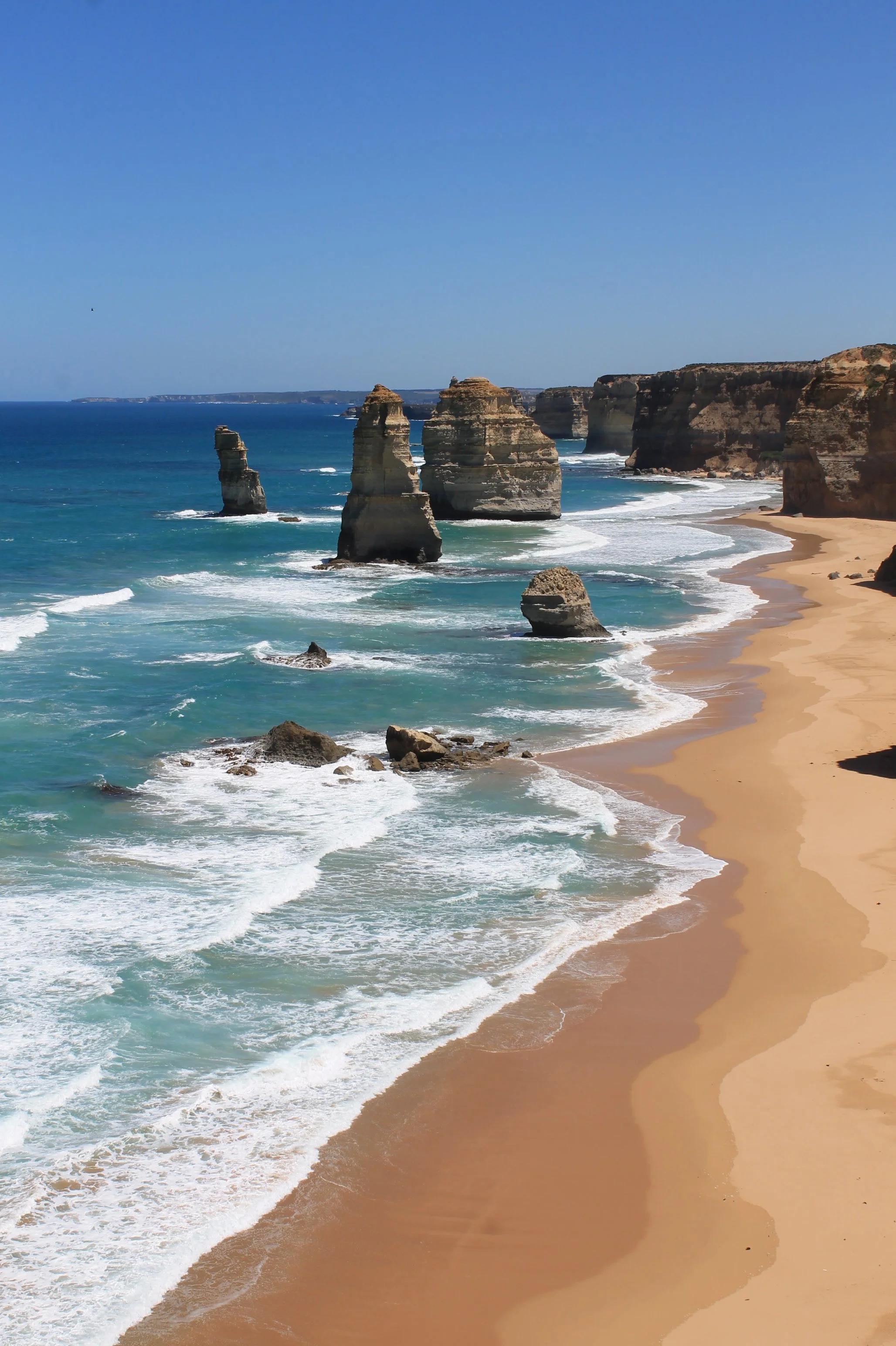 Photo of the Twelve Apostles, limestone stacks along the Great Ocean Road in Australia, with ocean waves crashing against the rocks and a sandy beach in the foreground under a clear blue sky.