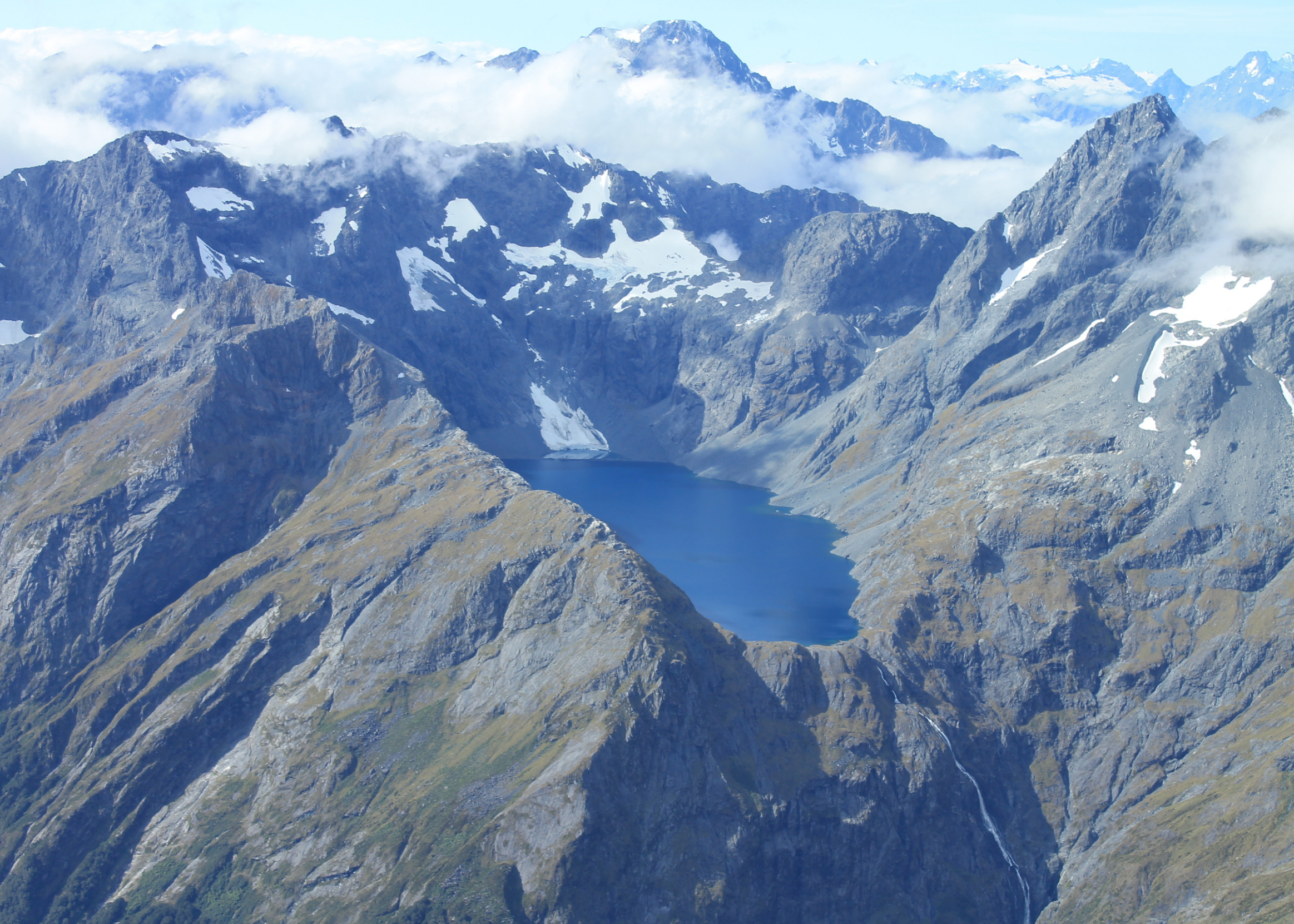 A scenic landscape of rugged mountain peaks with patches of snow, a partly cloudy sky, and a mountain lake below.