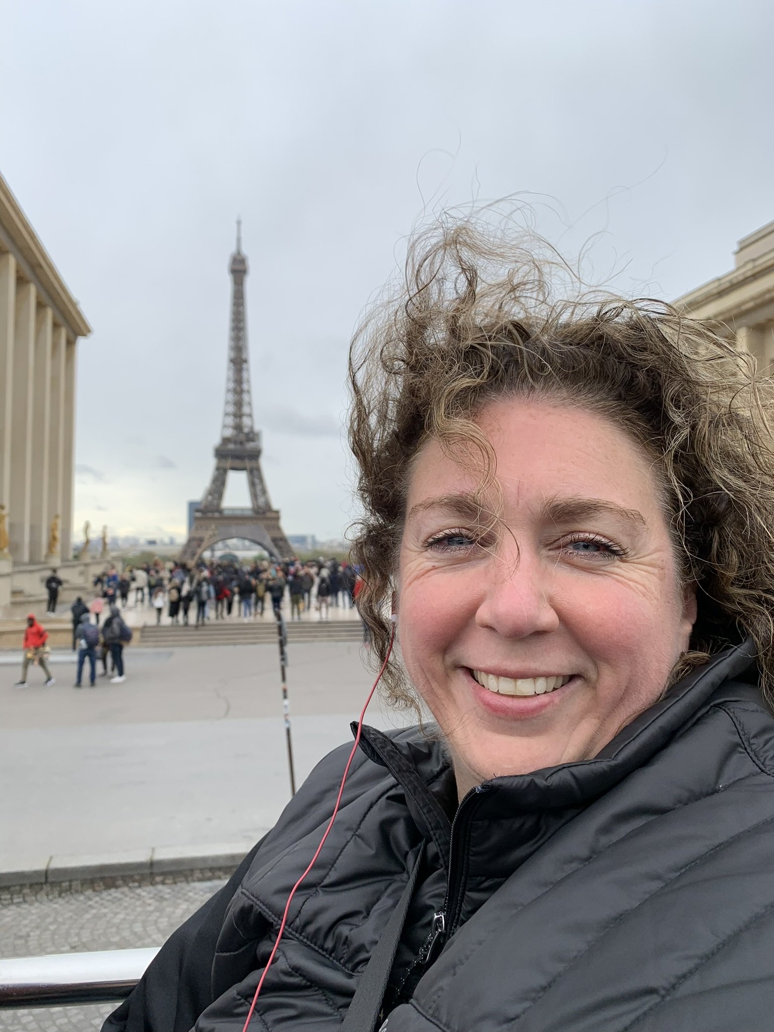 Smiling woman taking a selfie in front of the Eiffel Tower in Paris, France, with a crowd of tourists in the background.