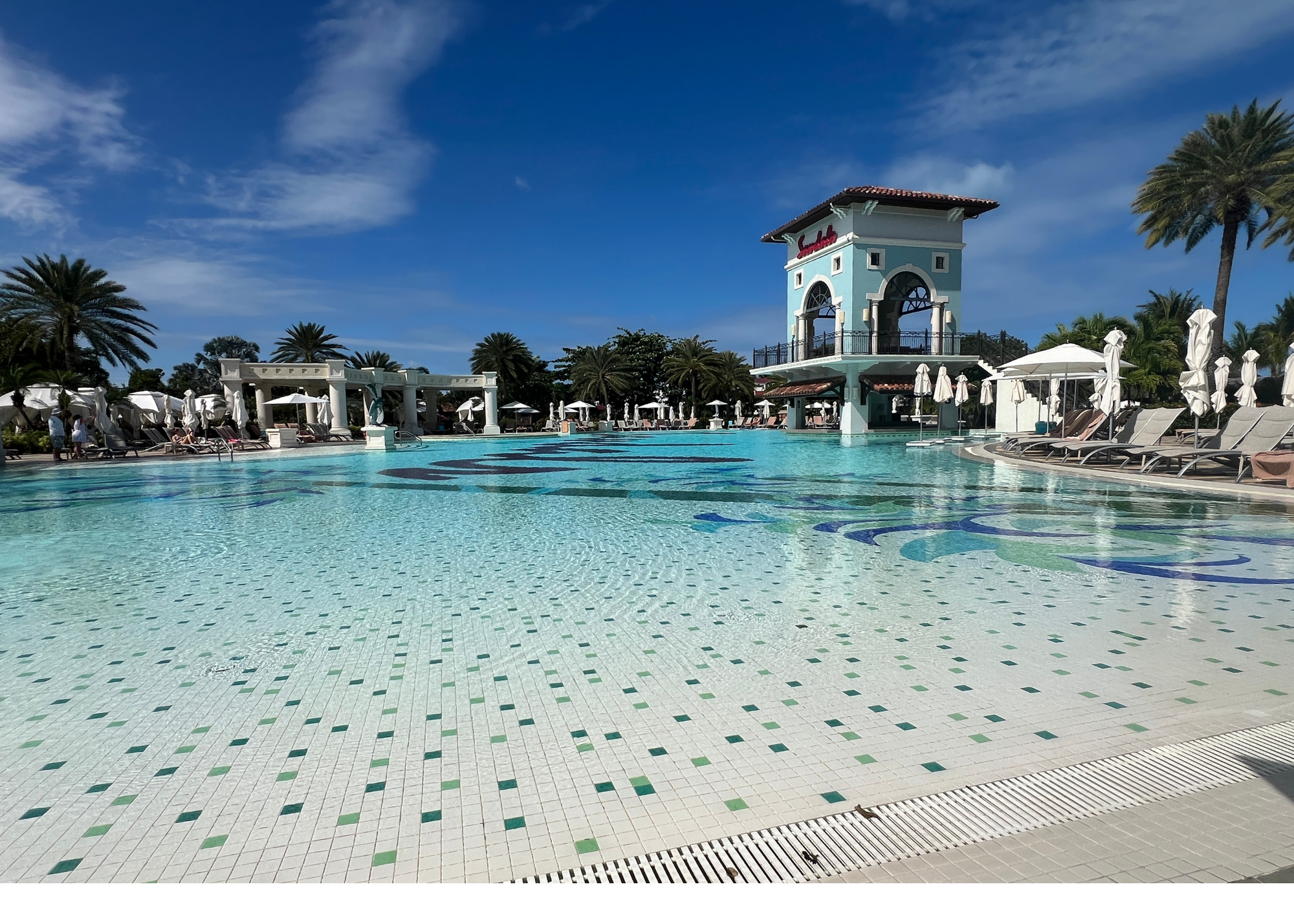 A large swimming pool with a tiled bottom, surrounded by lounge chairs and white umbrellas, with a blue sky and palm trees in the background, at a resort.